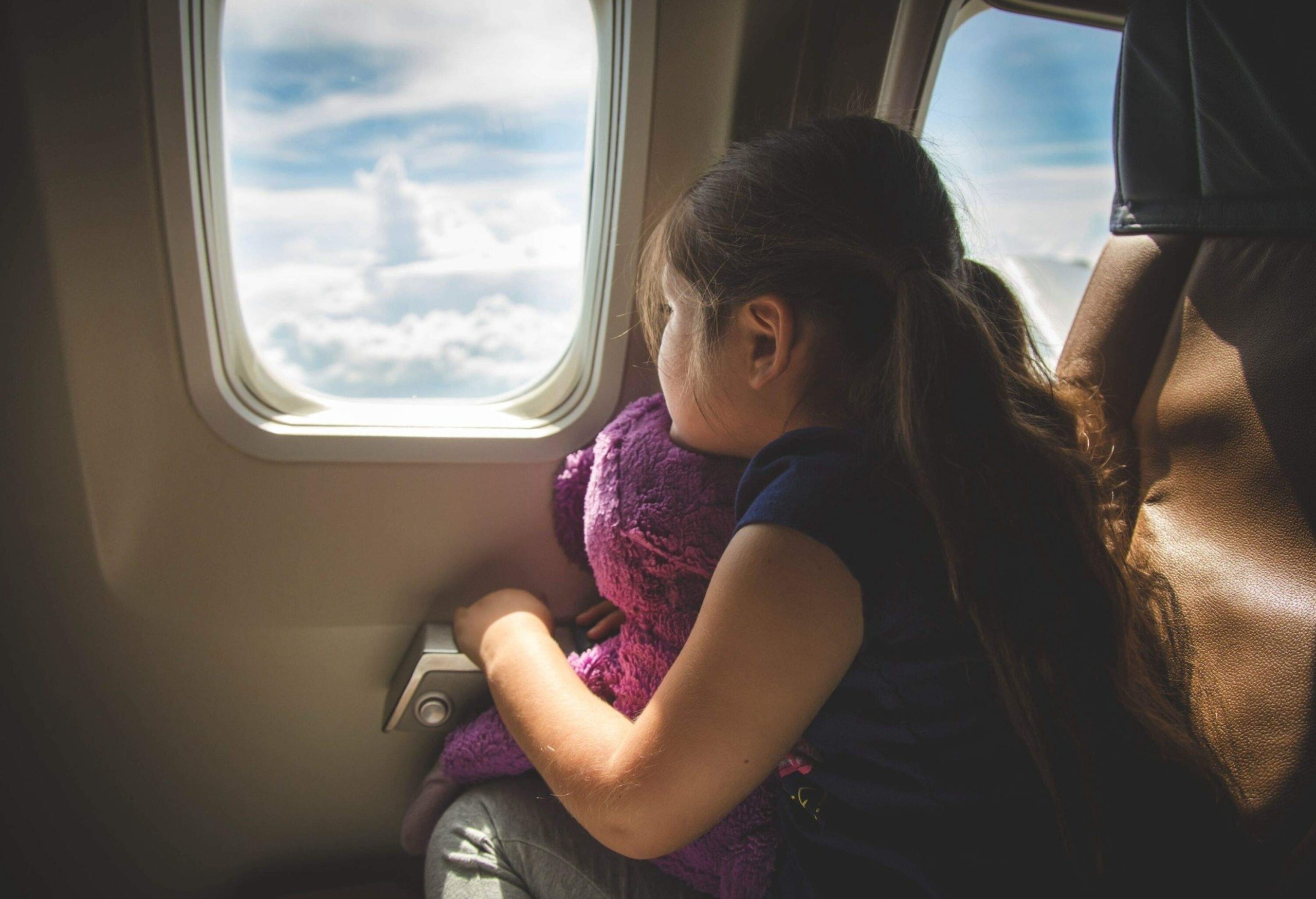 A little girl holding a purple stuffed toy as she looks out the window of an airplane.