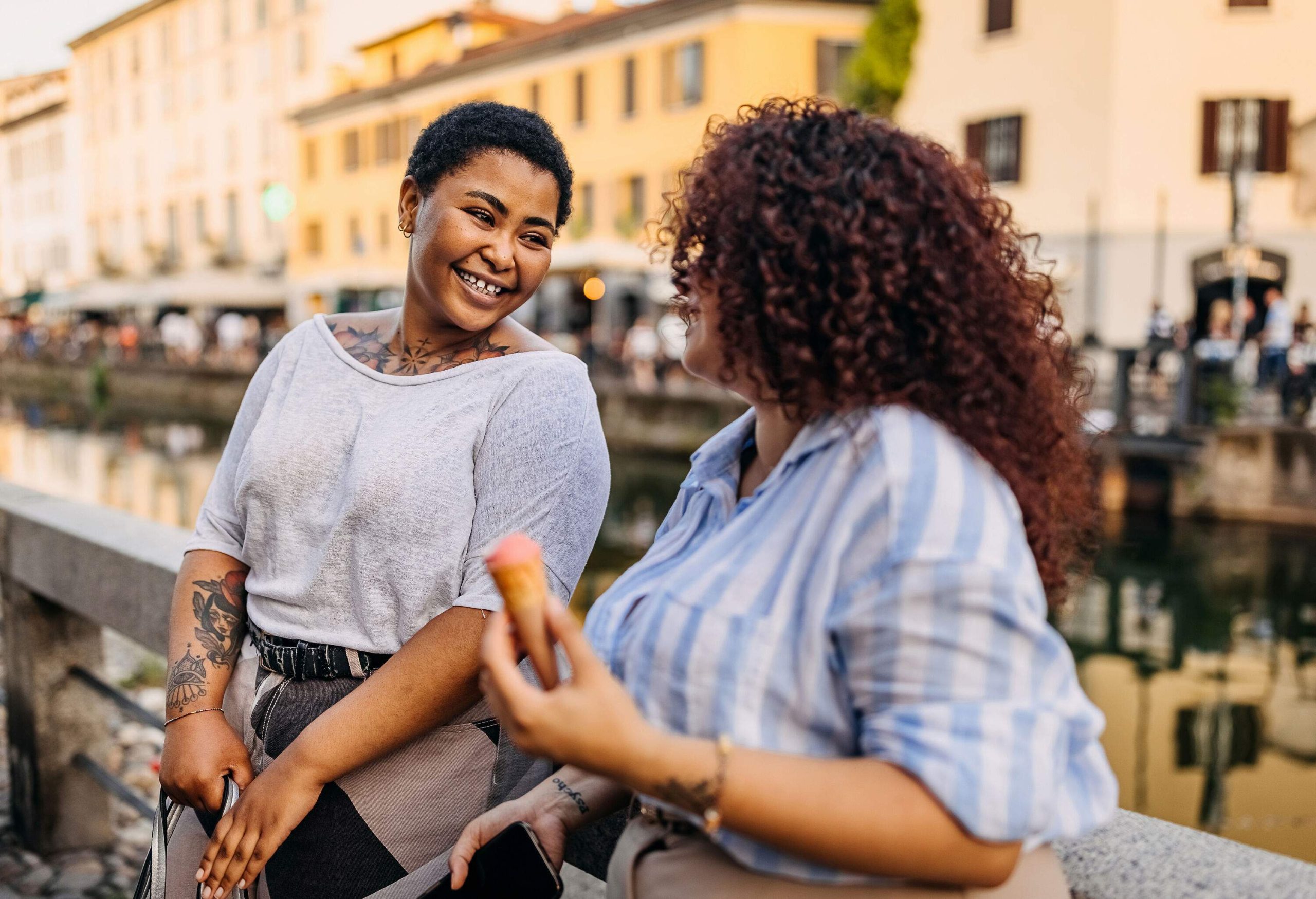 Two women engage in a relaxed conversation as they lean on a guardrail by the canal.