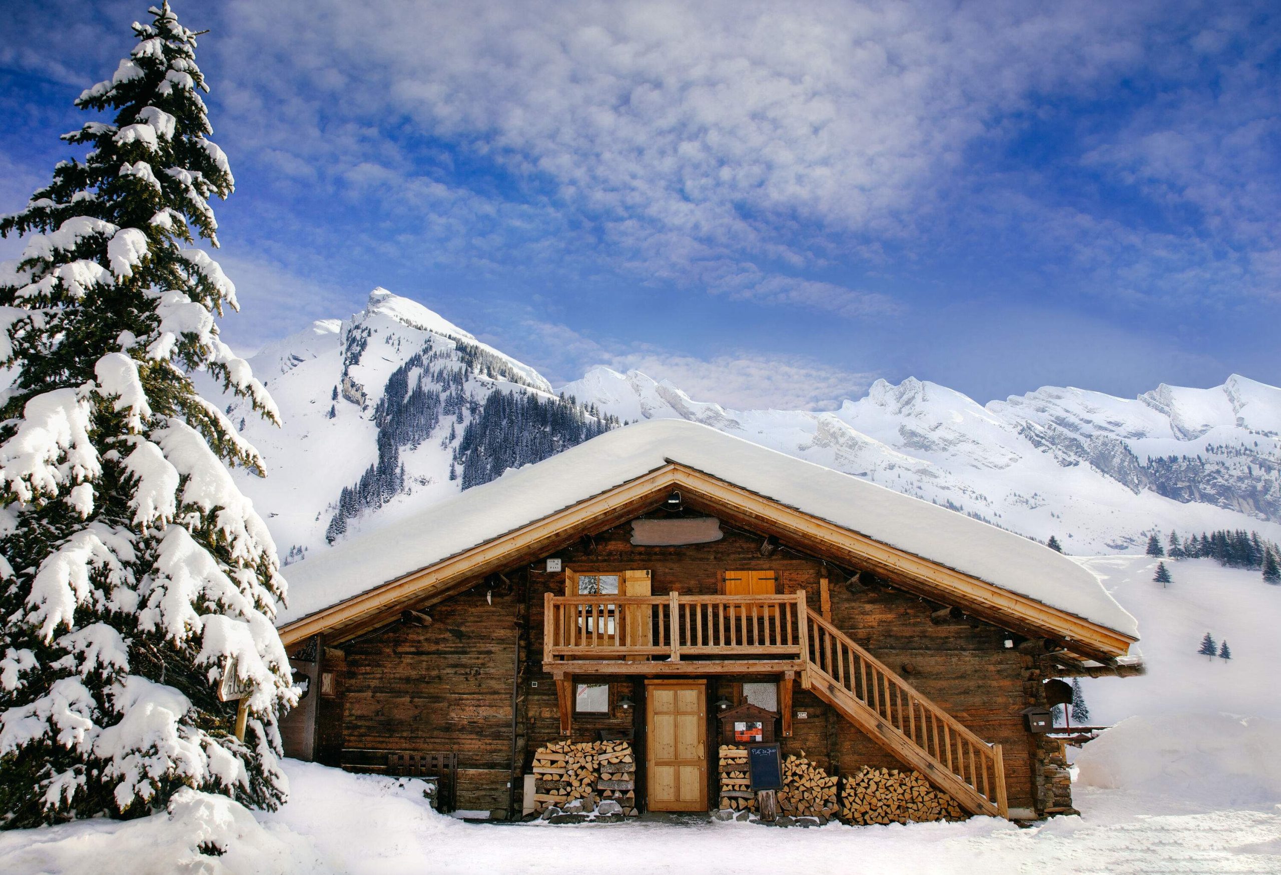 A wooden cabin with a roof covered in a thick layer of snow set in the mountains.