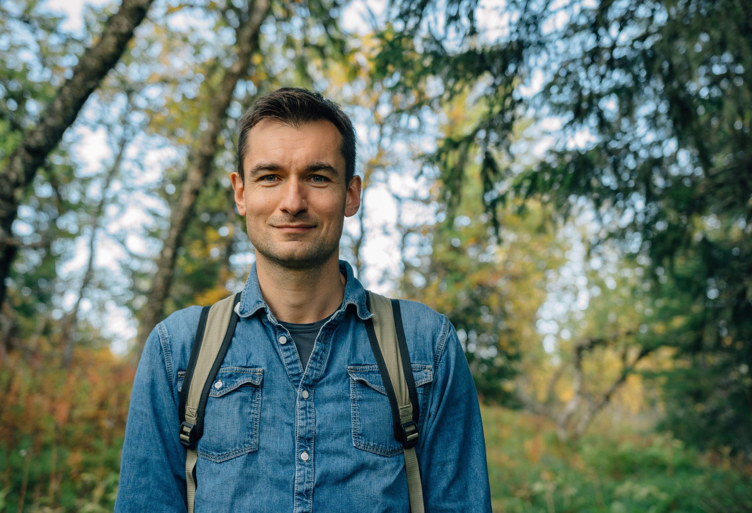 A portrait of a smiling man wearing a denim polo and a backpack against a nature background.