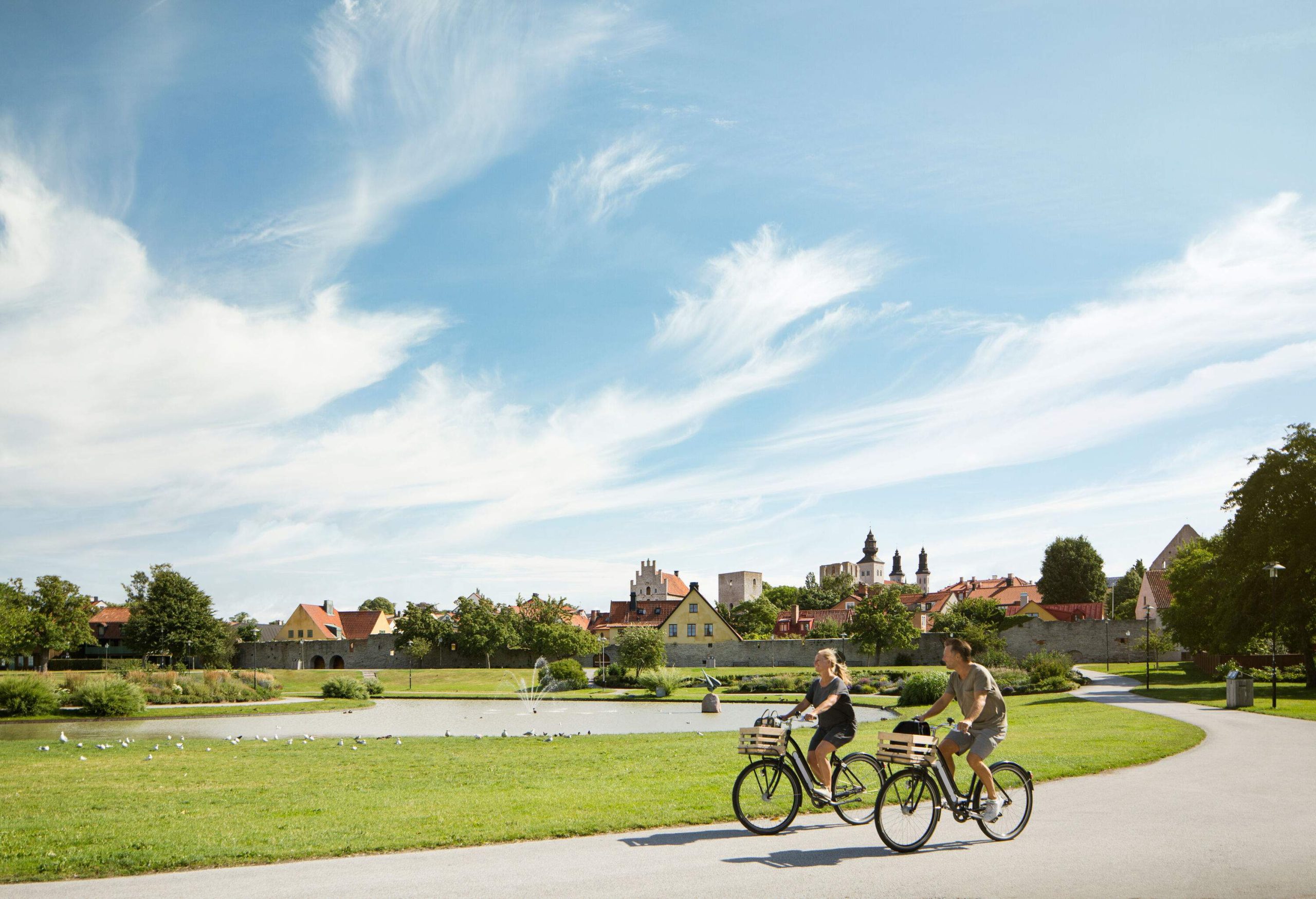 Two happy cyclists ride near a pond set in a lush lawn.