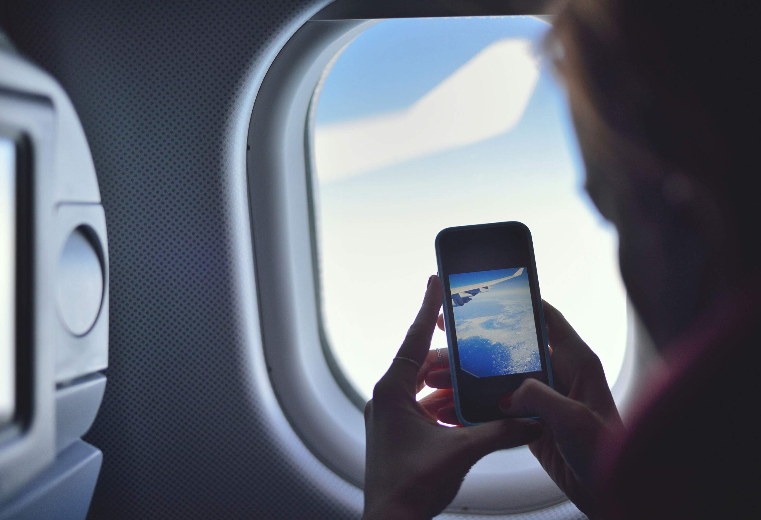 A passenger seated inside an aircraft by the window captures a photo of the white clouds using her smartphone.