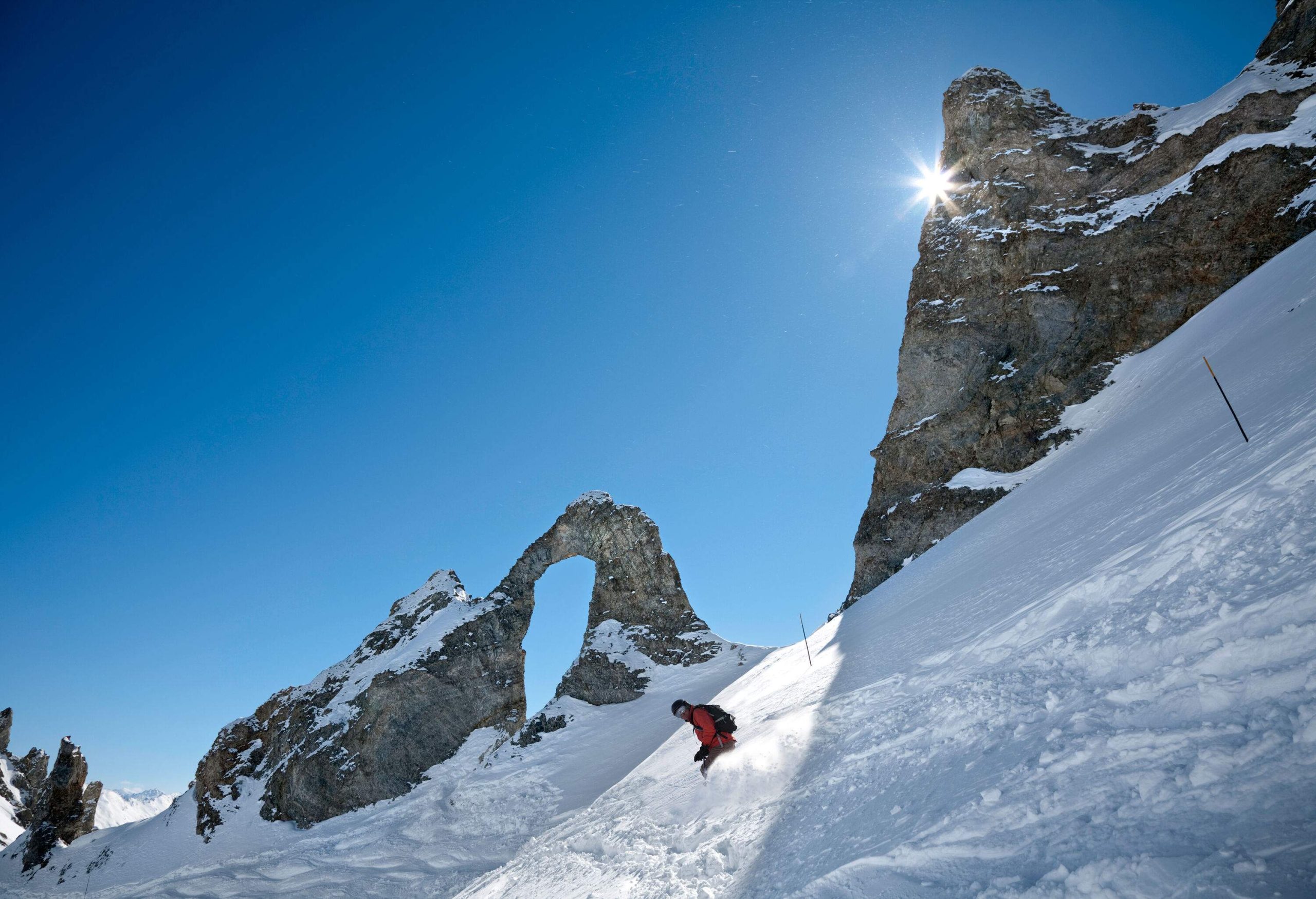 Skier going down a slope with the sun peeking through the mountains.