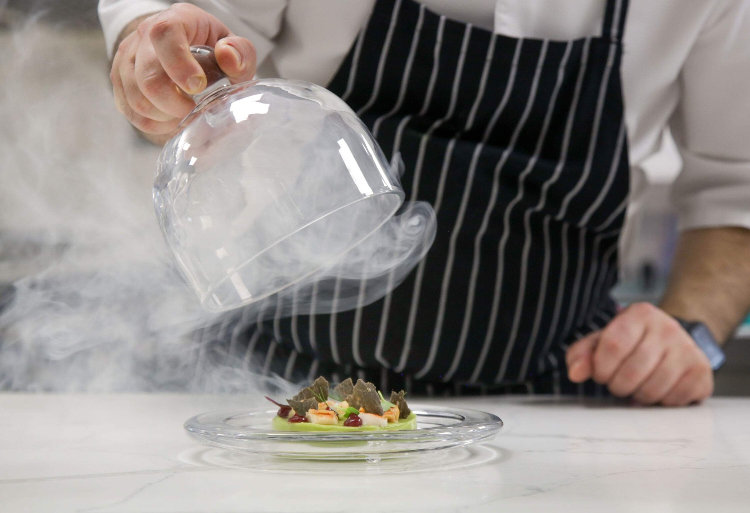 A hand lifts a glass lid of a smoky dessert on a plate.