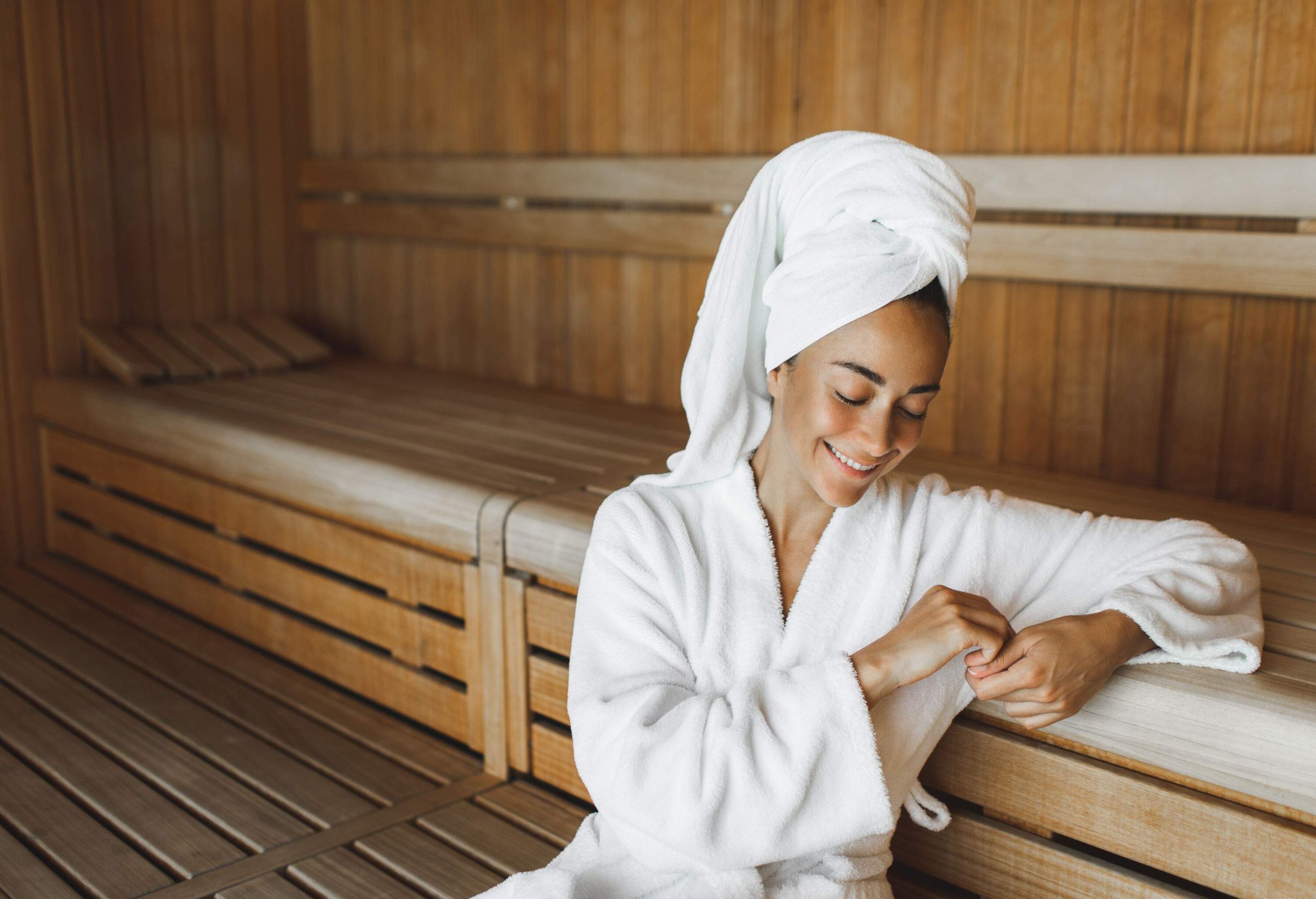 A woman in a bathrobe and towel on her head smiles as she sits inside a dry sauna.