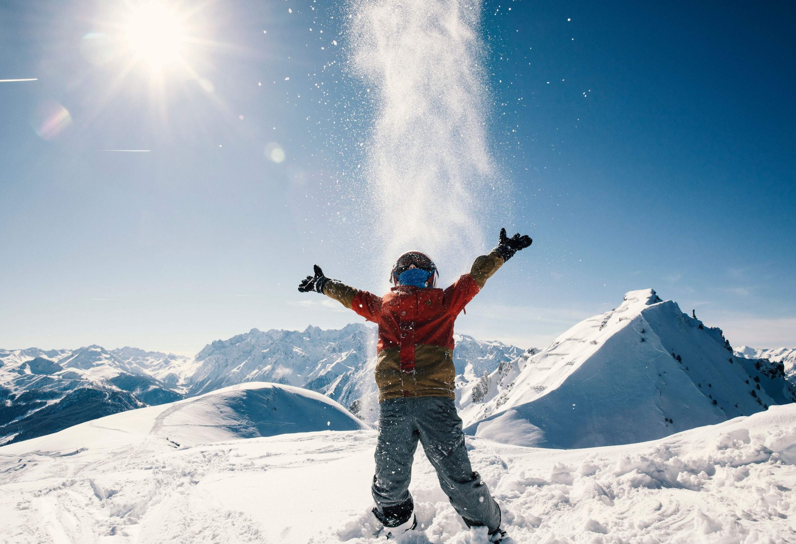 A young boy on a hilltop throwing snow in the air under bright sunlight.