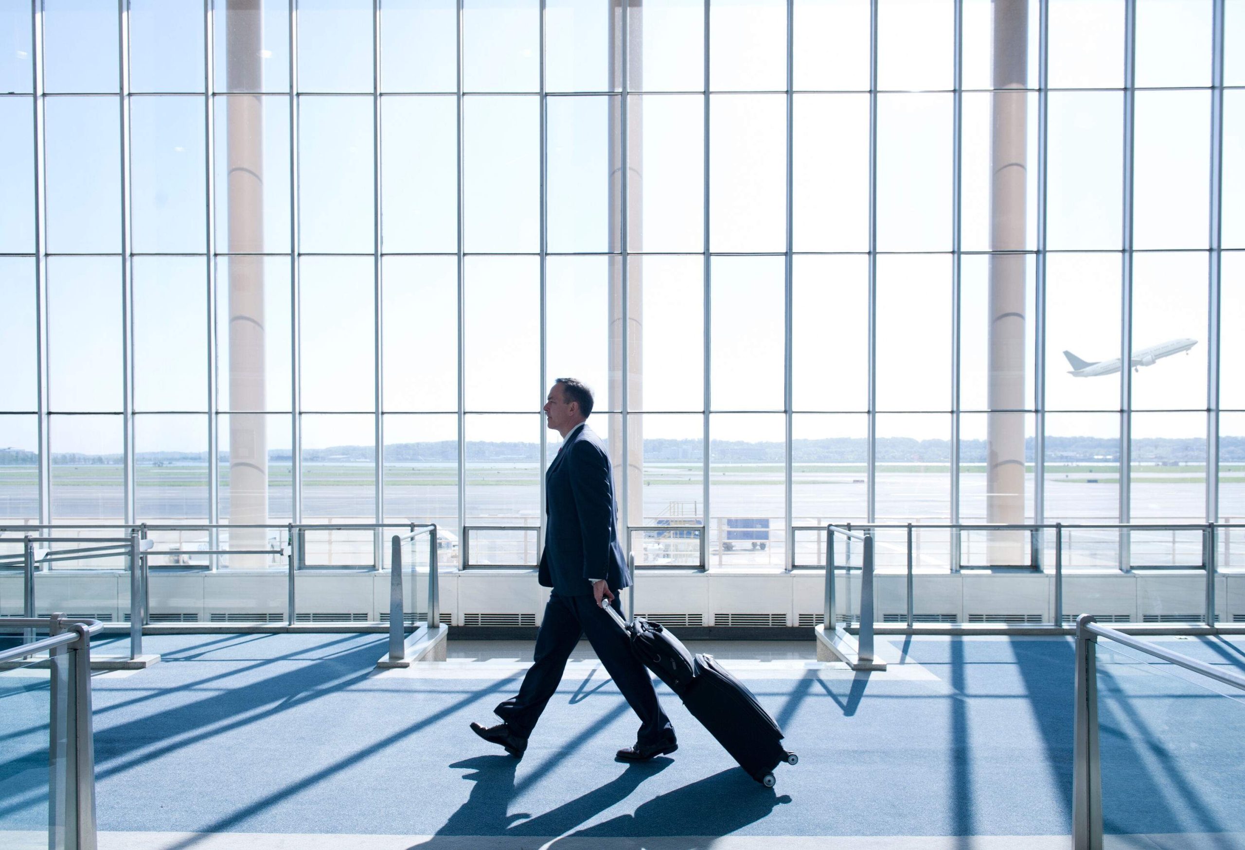 A professional traveller, confidently navigating the airport with luggage in tow, catches a glimpse of a departing airplane through a glass wall.