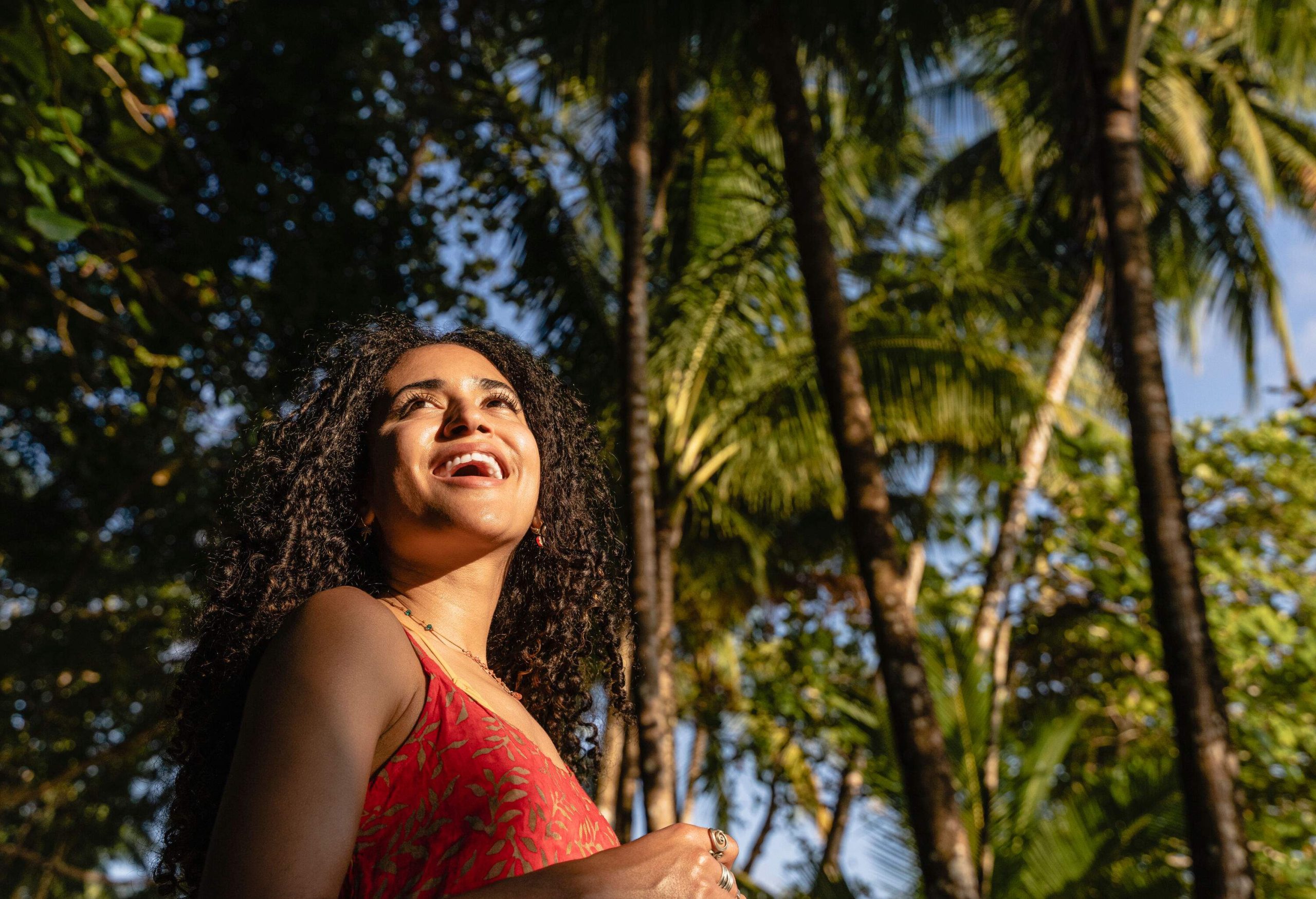 A curly-haired woman smiles under a canopy of palm trees.