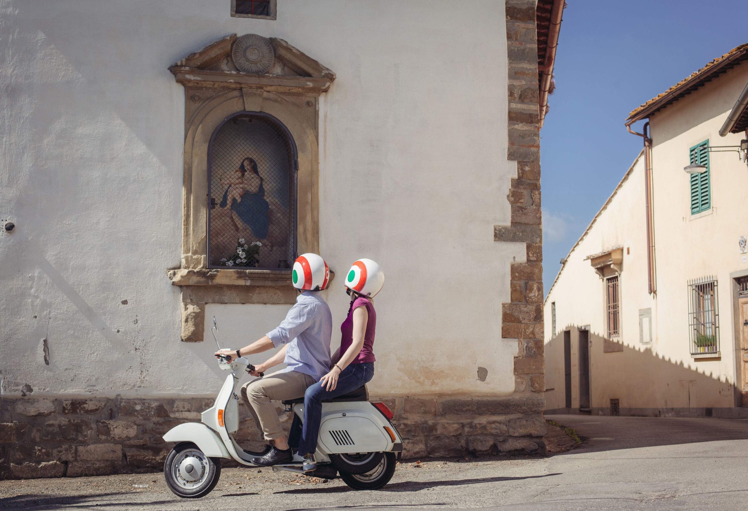 Two individuals on a classic white scooter are looking at a painting in the window of a building.