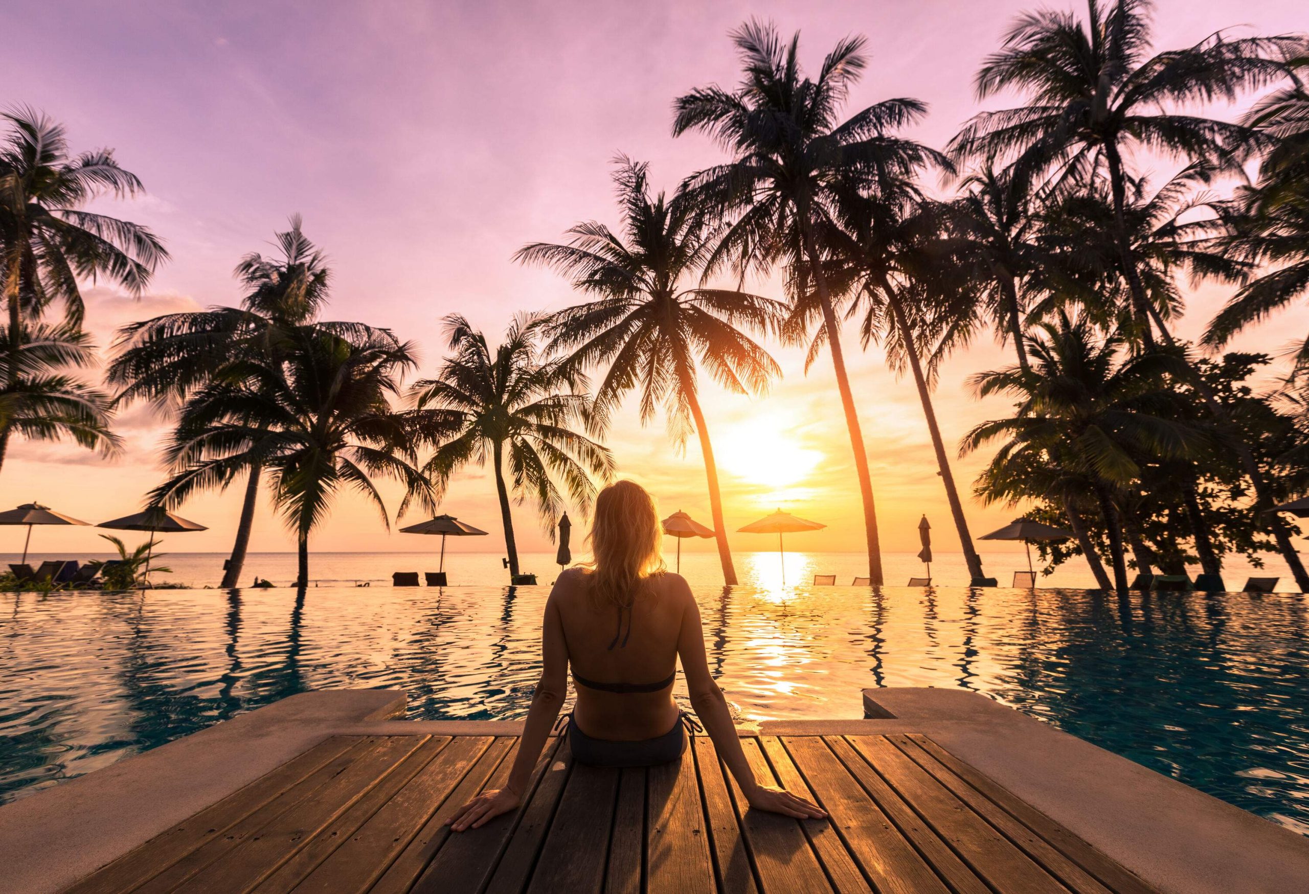 A woman sits on the deck of an infinity pool overlooking the tall palm trees on the beach against the scenic sunset.