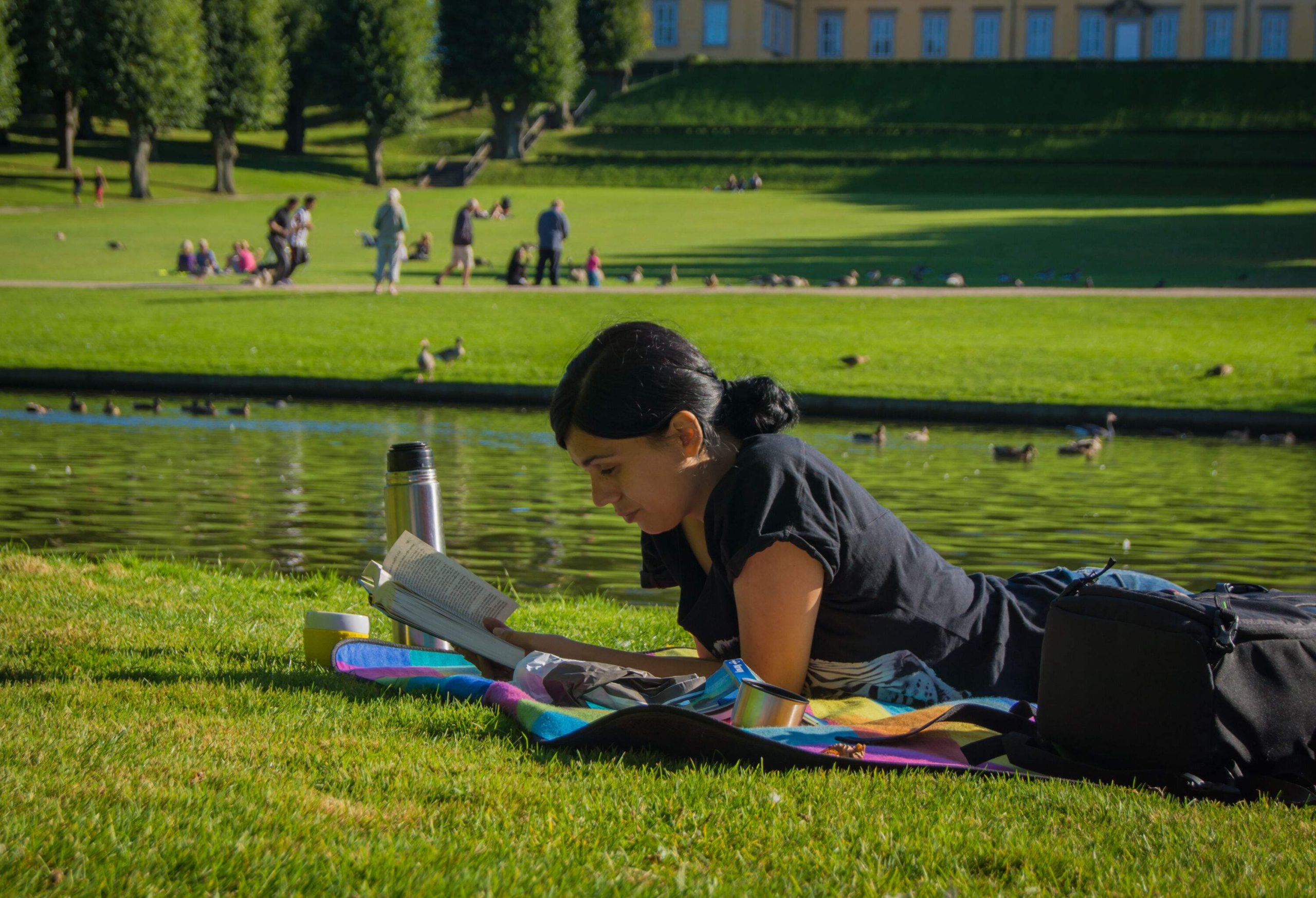 A young woman reading a book while lying on her stomach on the grass beside a pond.