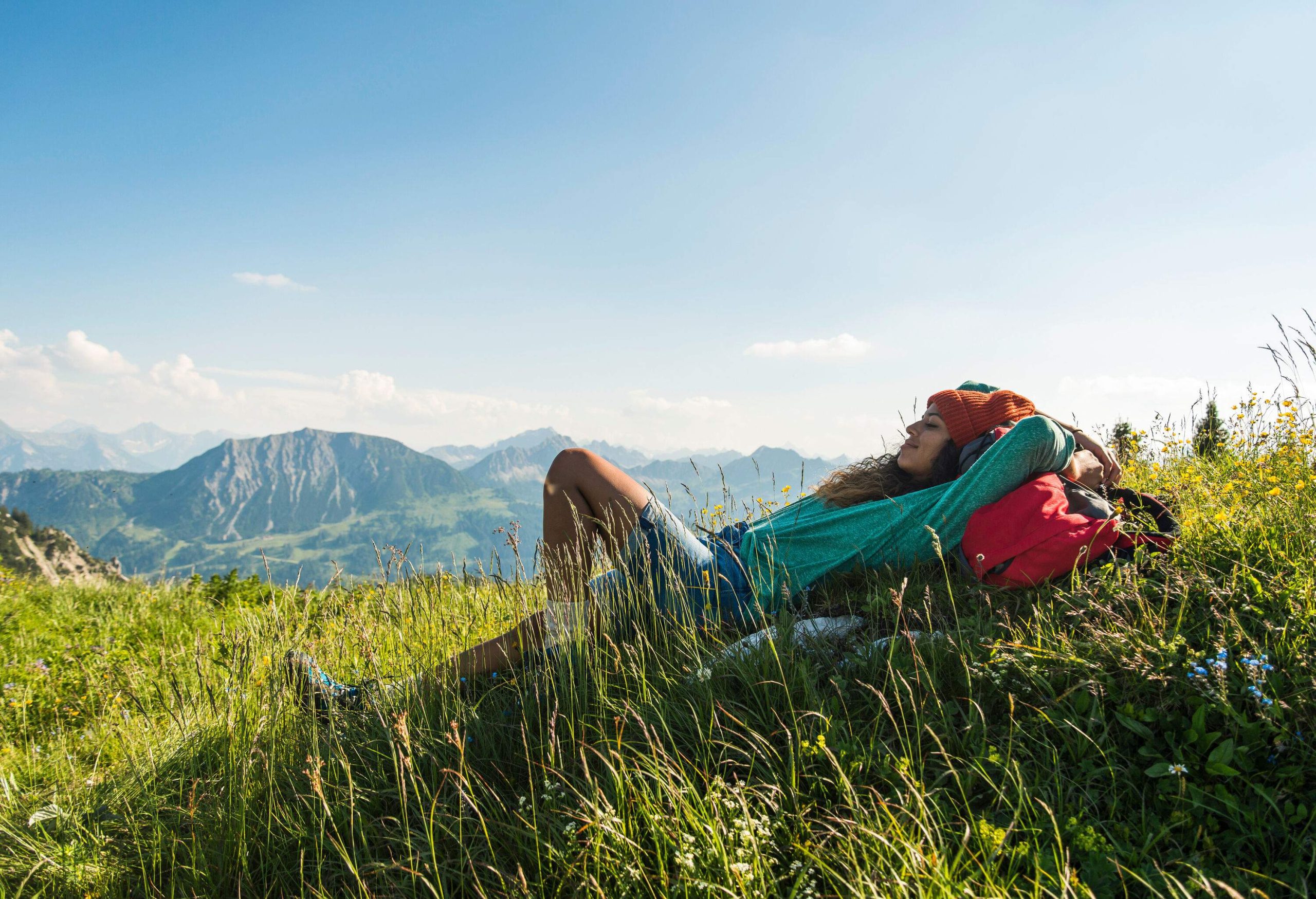 A woman lies on the grassy summit overlooking the mountain range against the cloudy blue sky.