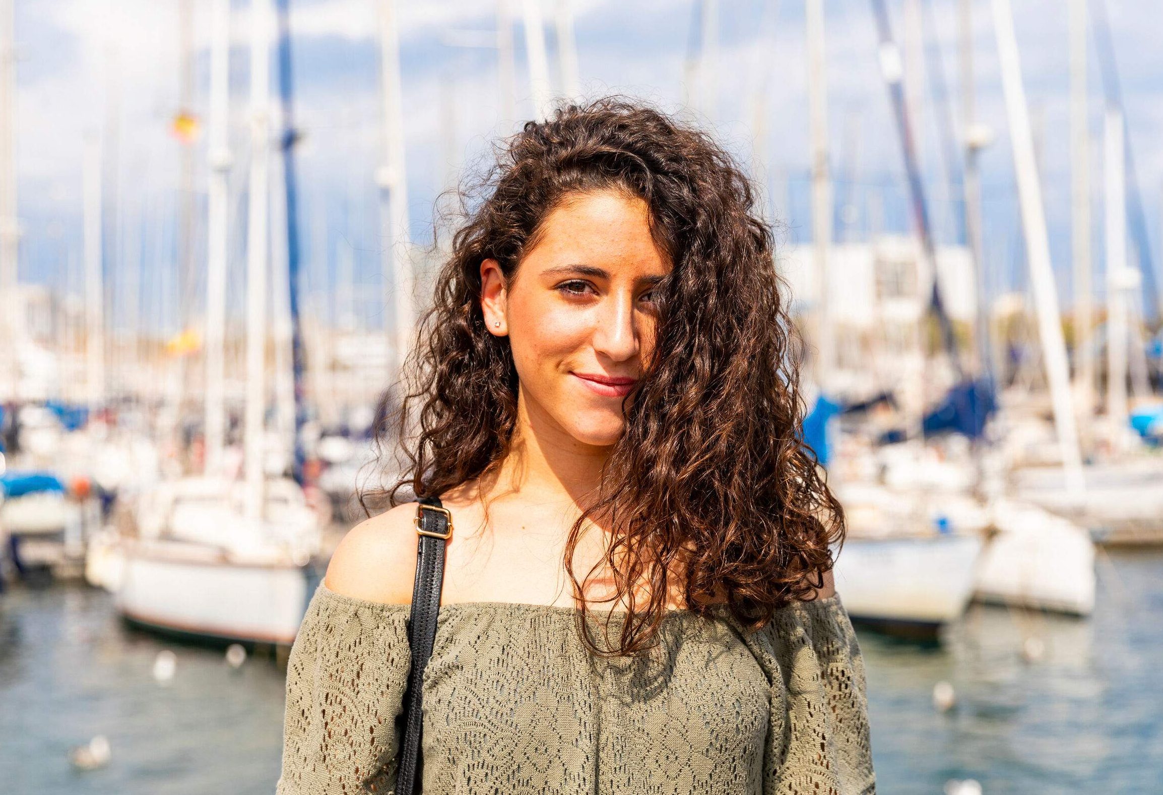 A curly-haired woman smiles against the backdrop of yachts docked on a harbour.