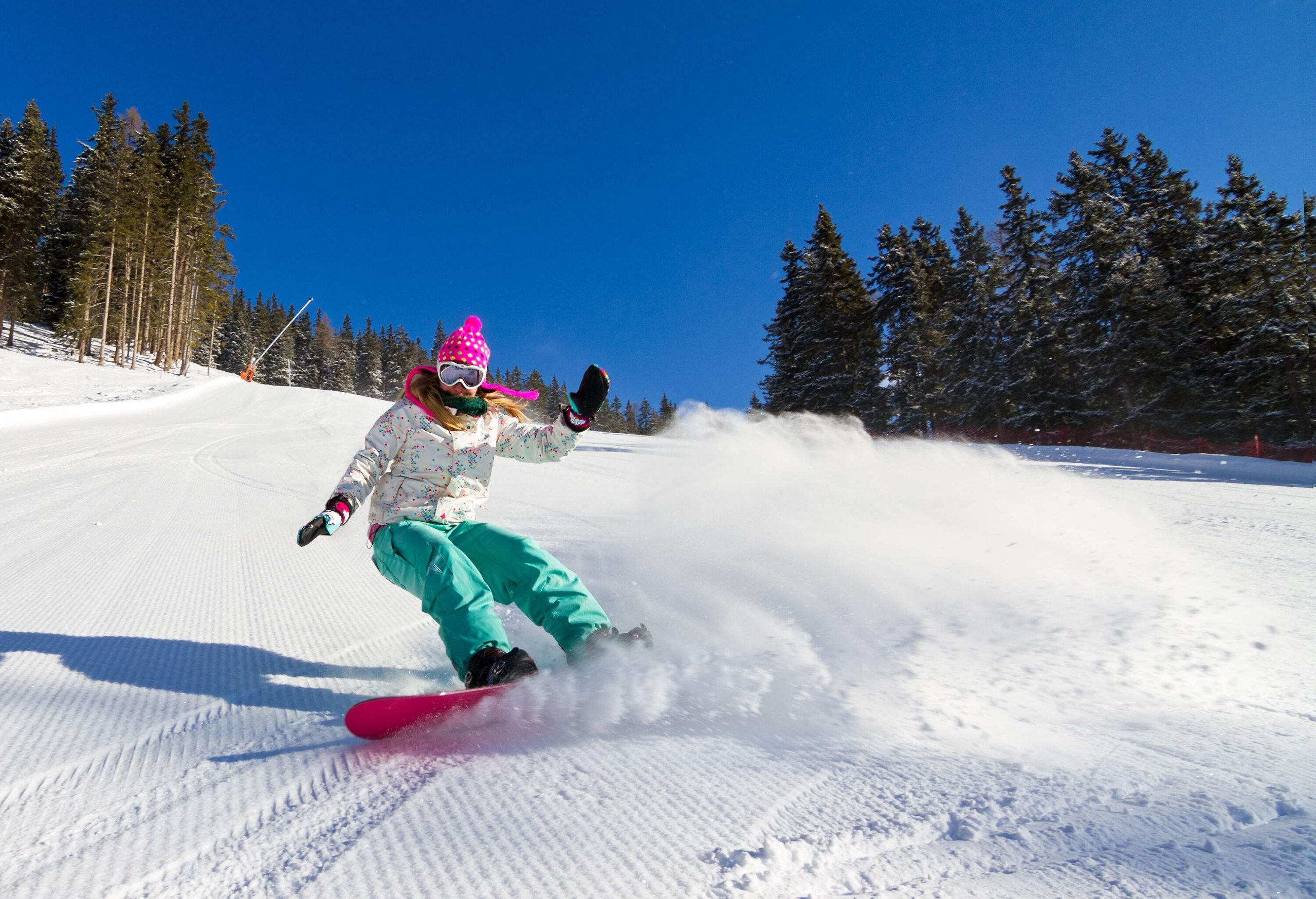 A woman on a pink snowboard making a skidded turn on a well-groomed slope.