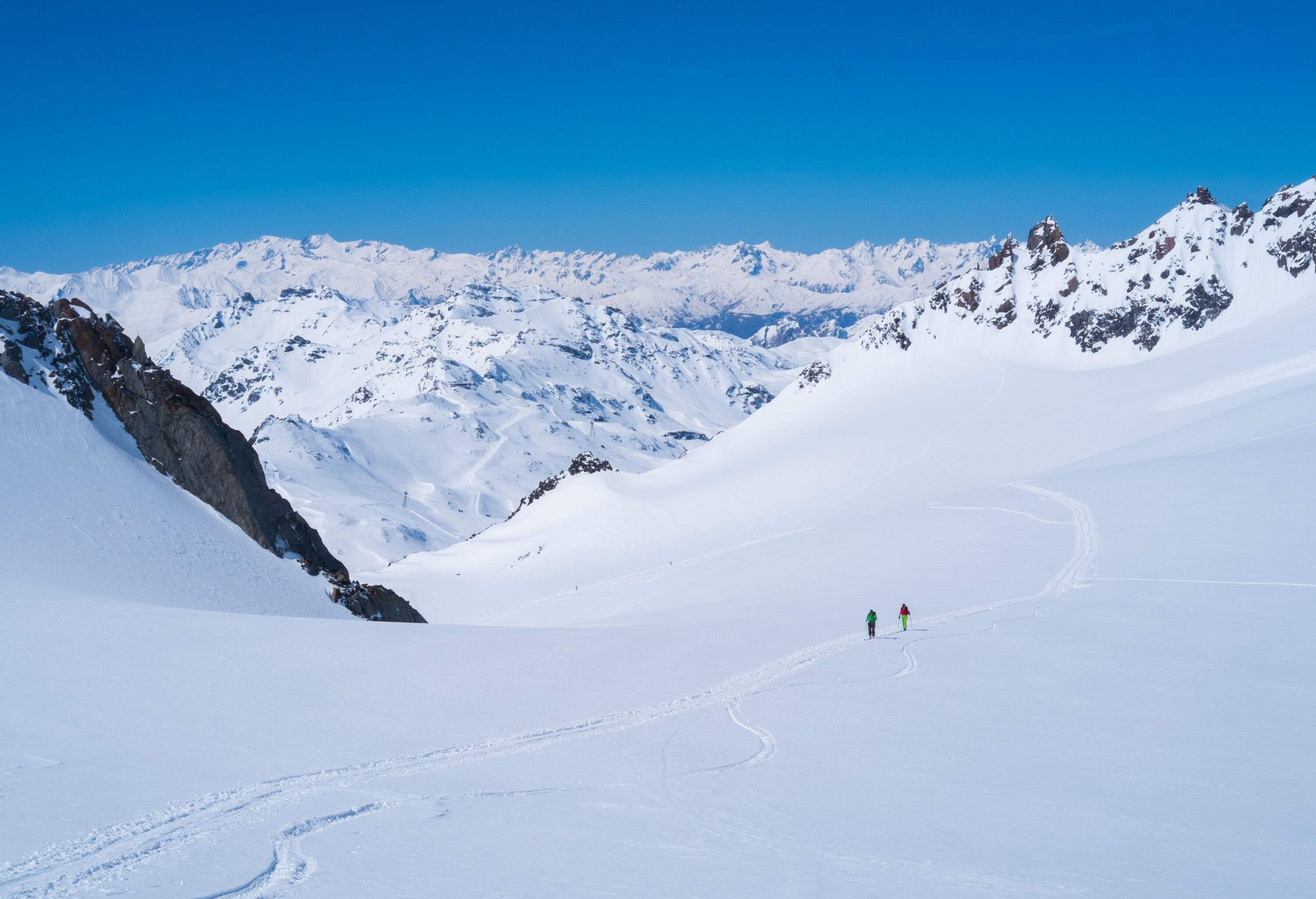 Two people walking across a large snowfield with views of snow-capped mountains on the horizon.
