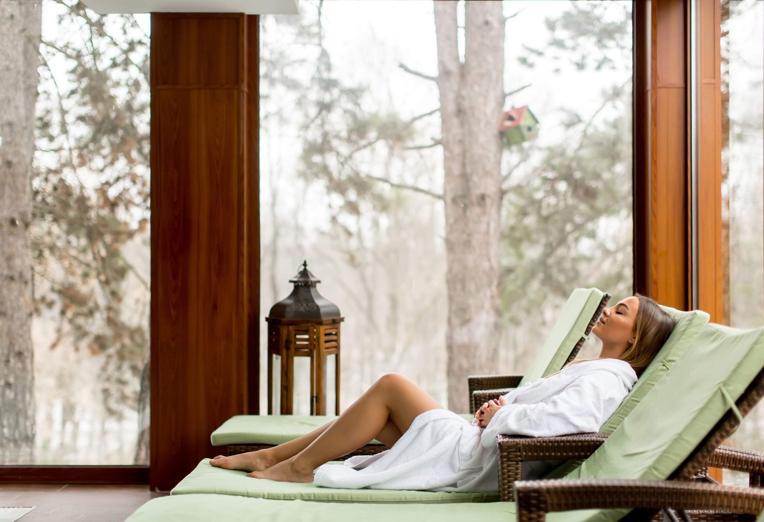 A woman in a white bathrobe rests on a reclining chair beside a big glass window with nature views.