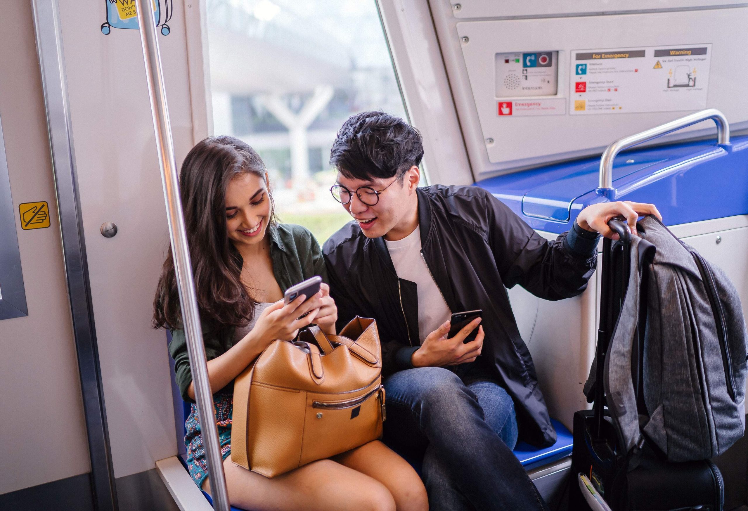 A couple smile as they sit in a train and look at a smartphone together.