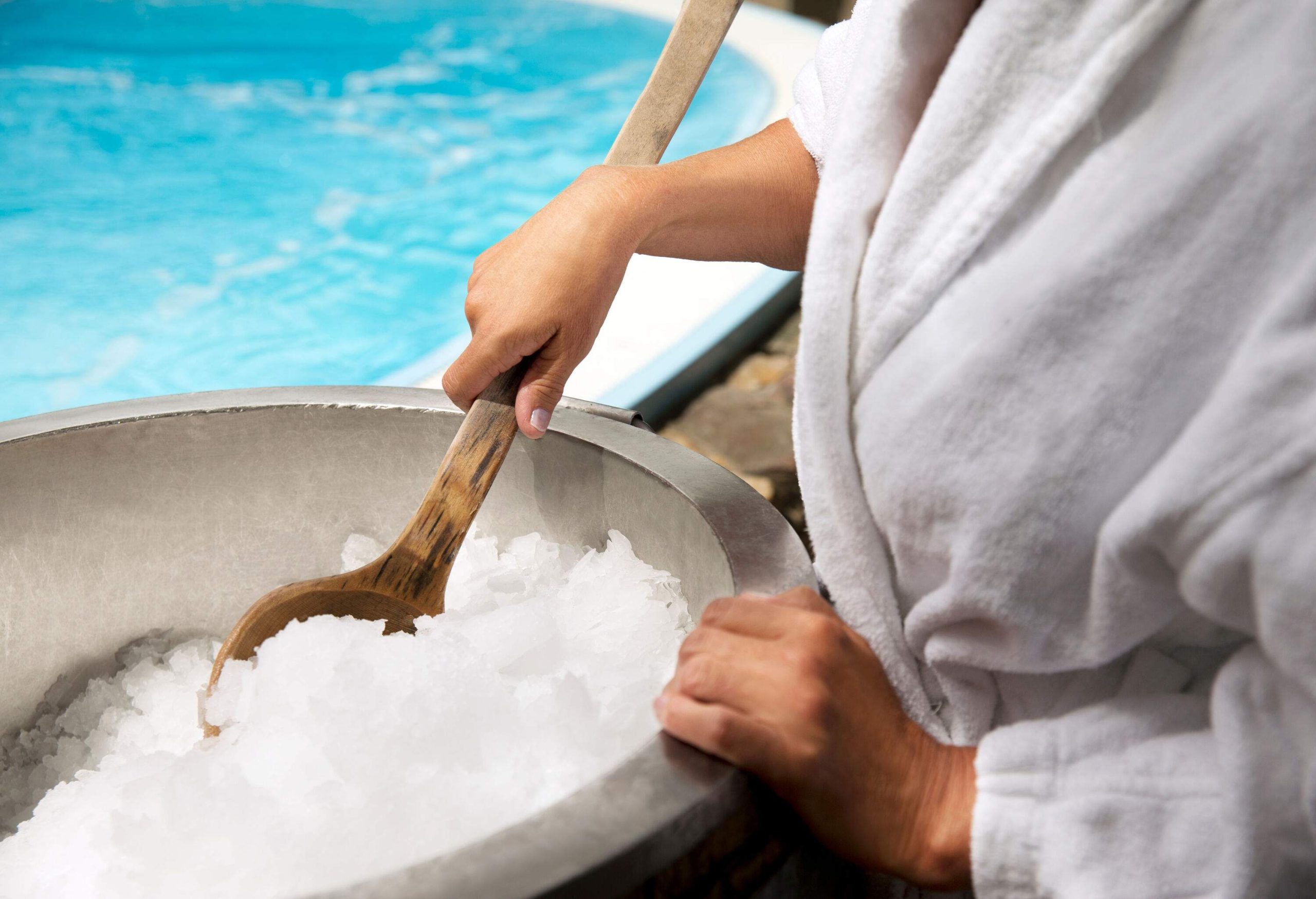 A hand scooping ice from a bucket using a wooden spoon on a poolside.