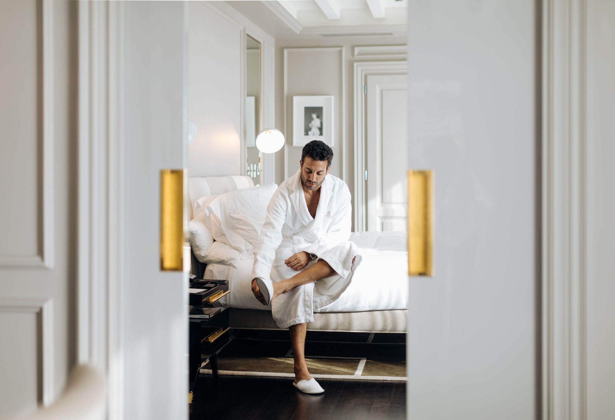 A man in a white robe sits in the bed of a luxury suite.