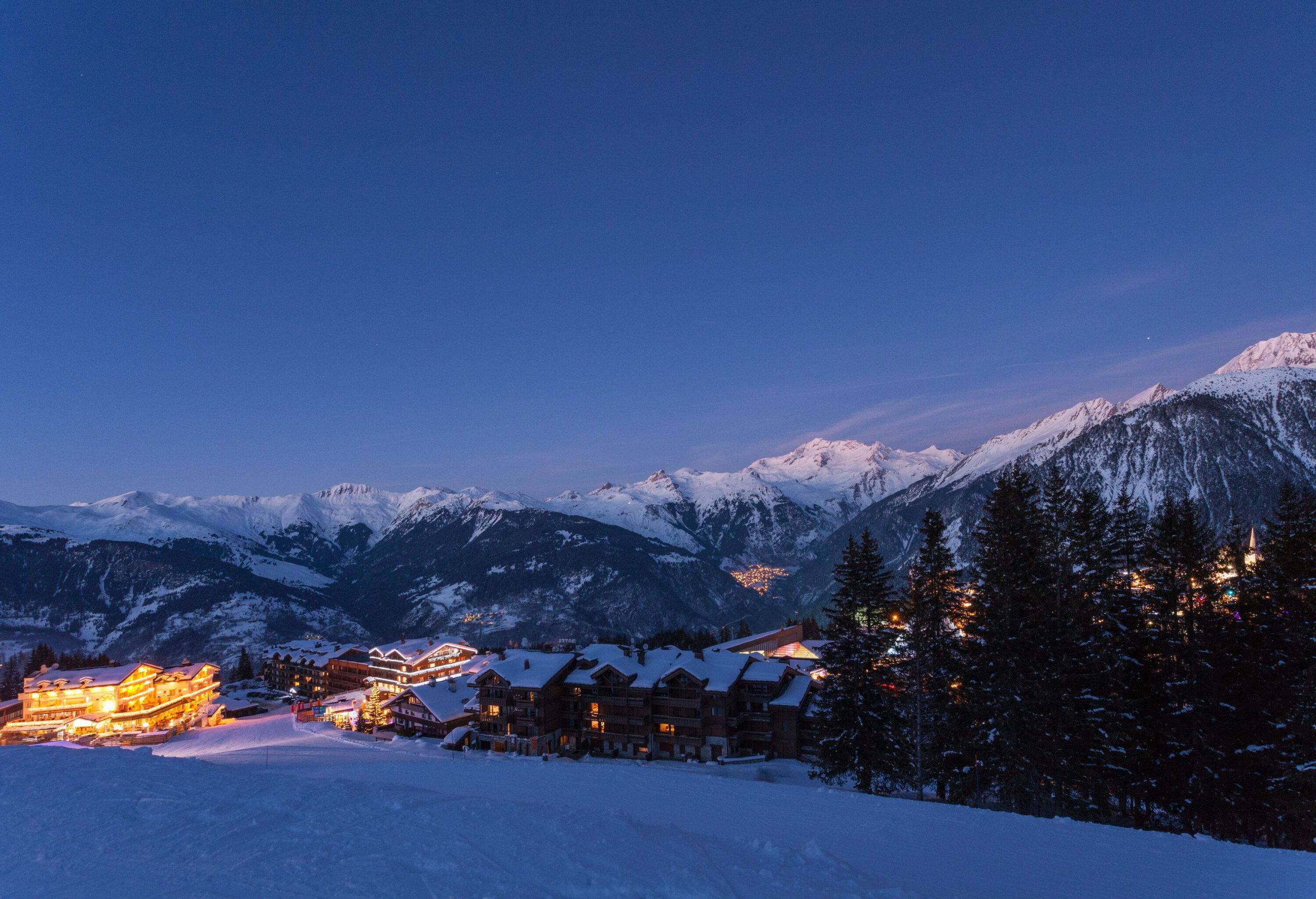 A settlement blanketed in snow and illuminated at night, surrounded by mountains and trees.