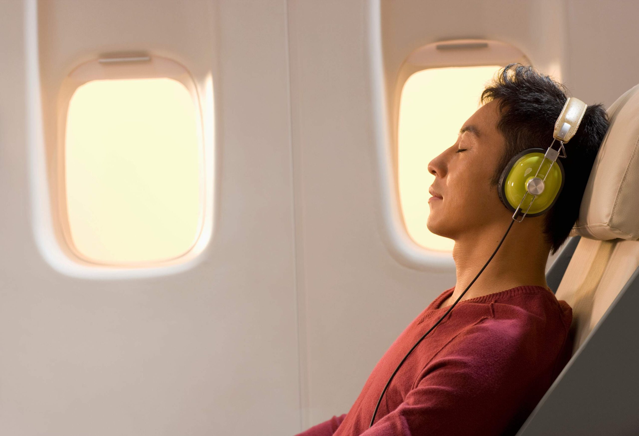 A male passenger in a window seat falling into slumber with headphones over his ears during a flight.