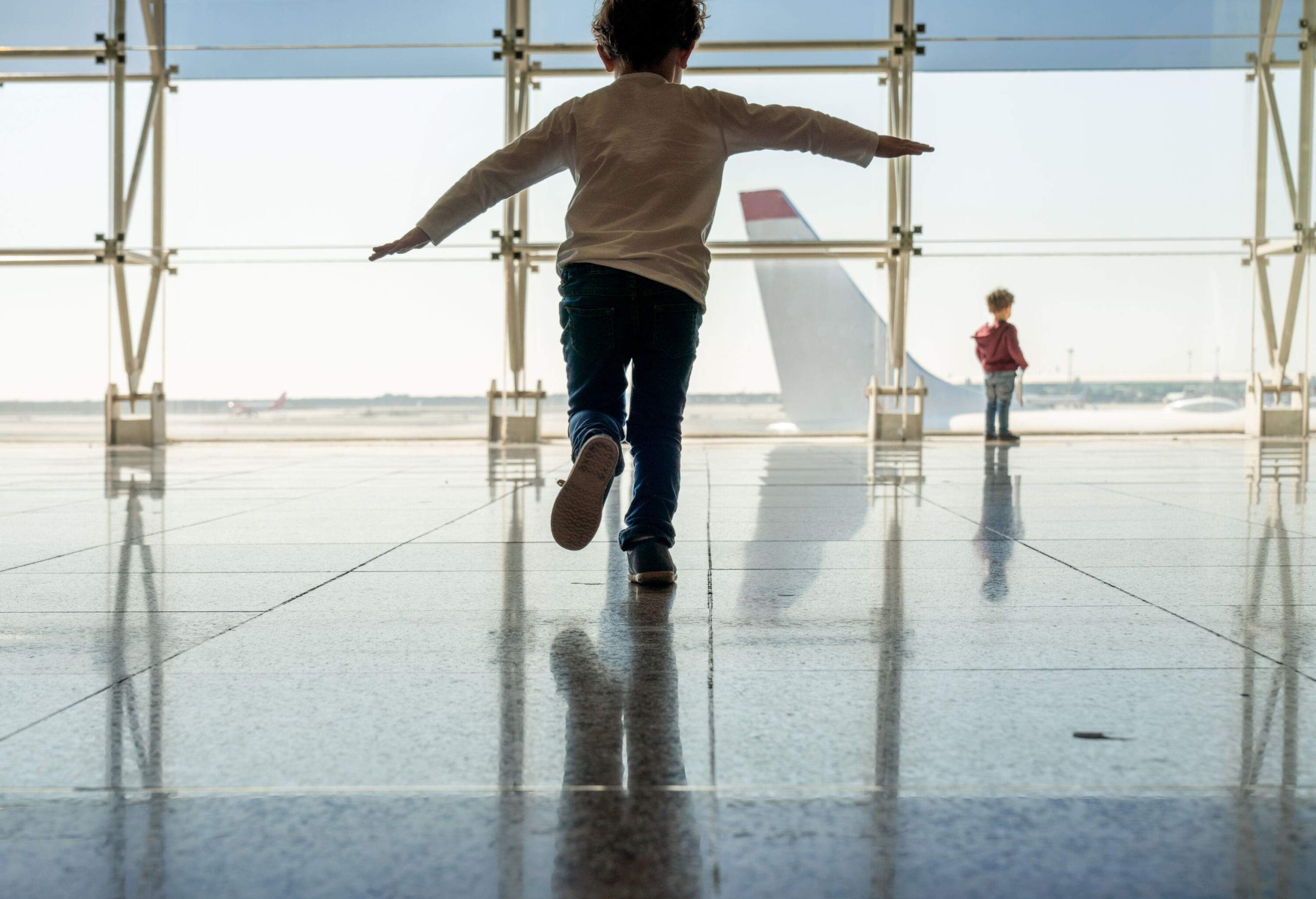 A boy balancing on one foot and another on the glass window looking at the airplane in the airport.