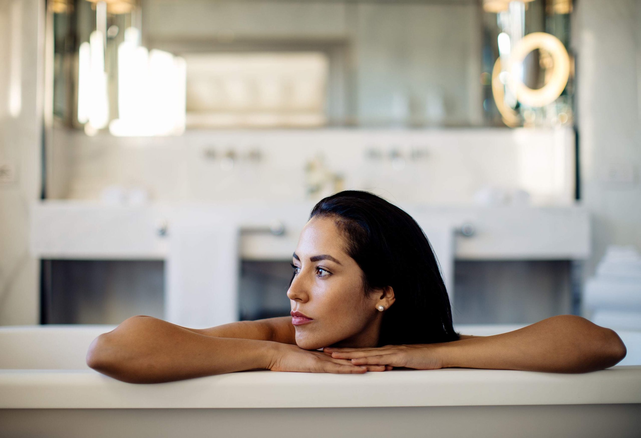 A woman submerged in a bathtub inside a brightly lit bathroom, looking to her right side.