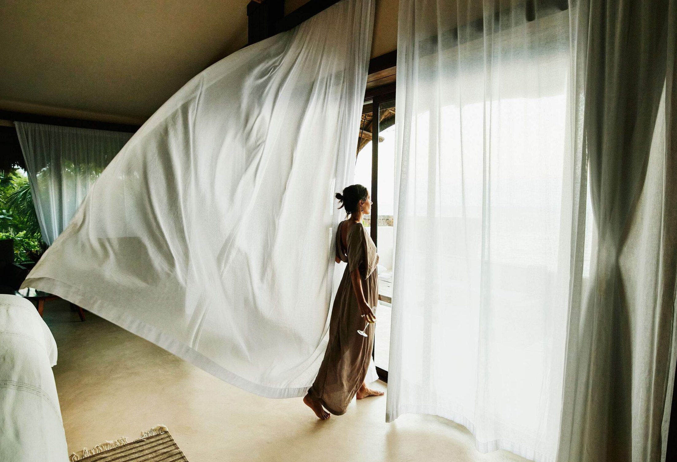 A woman stands beside a big window as the breeze flows through the curtains.