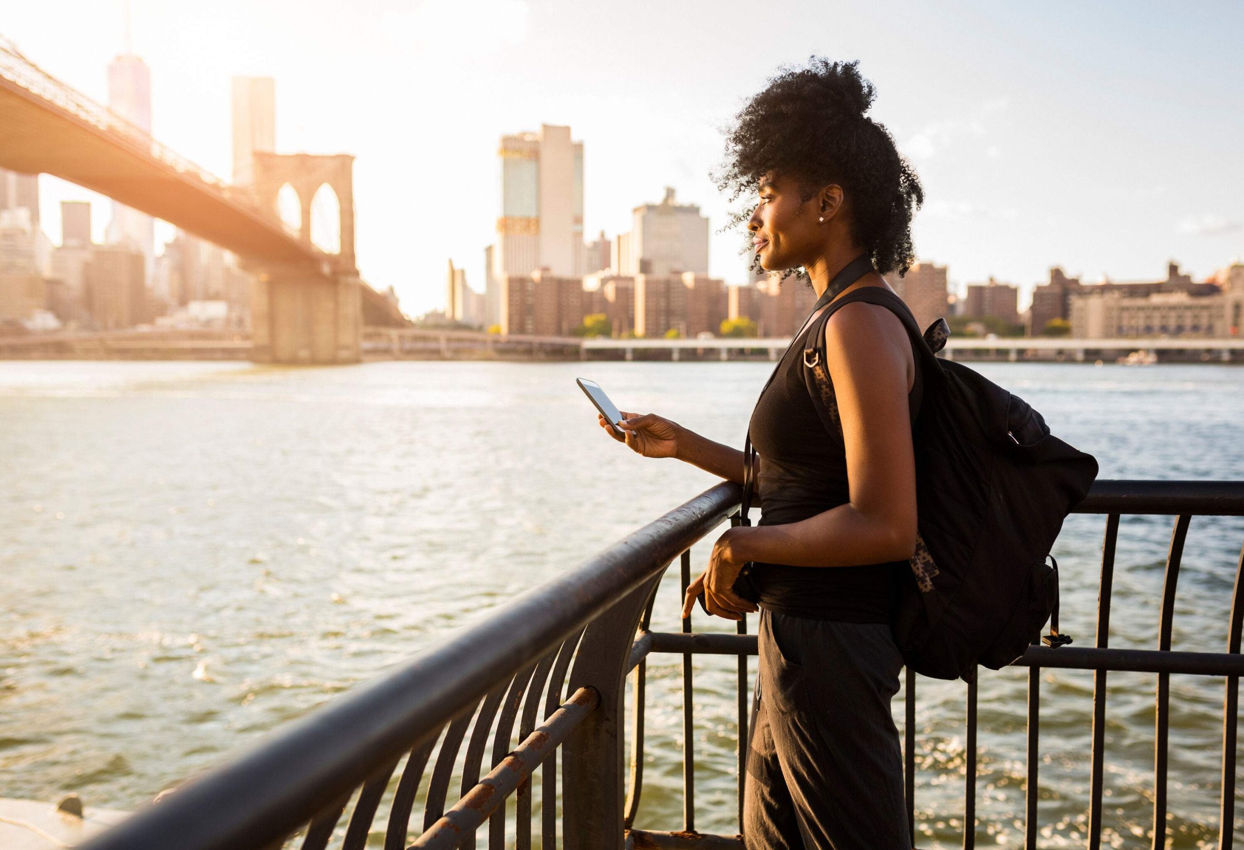 A curly-haired woman stands on a riverside railing and smiles while holding a phone.