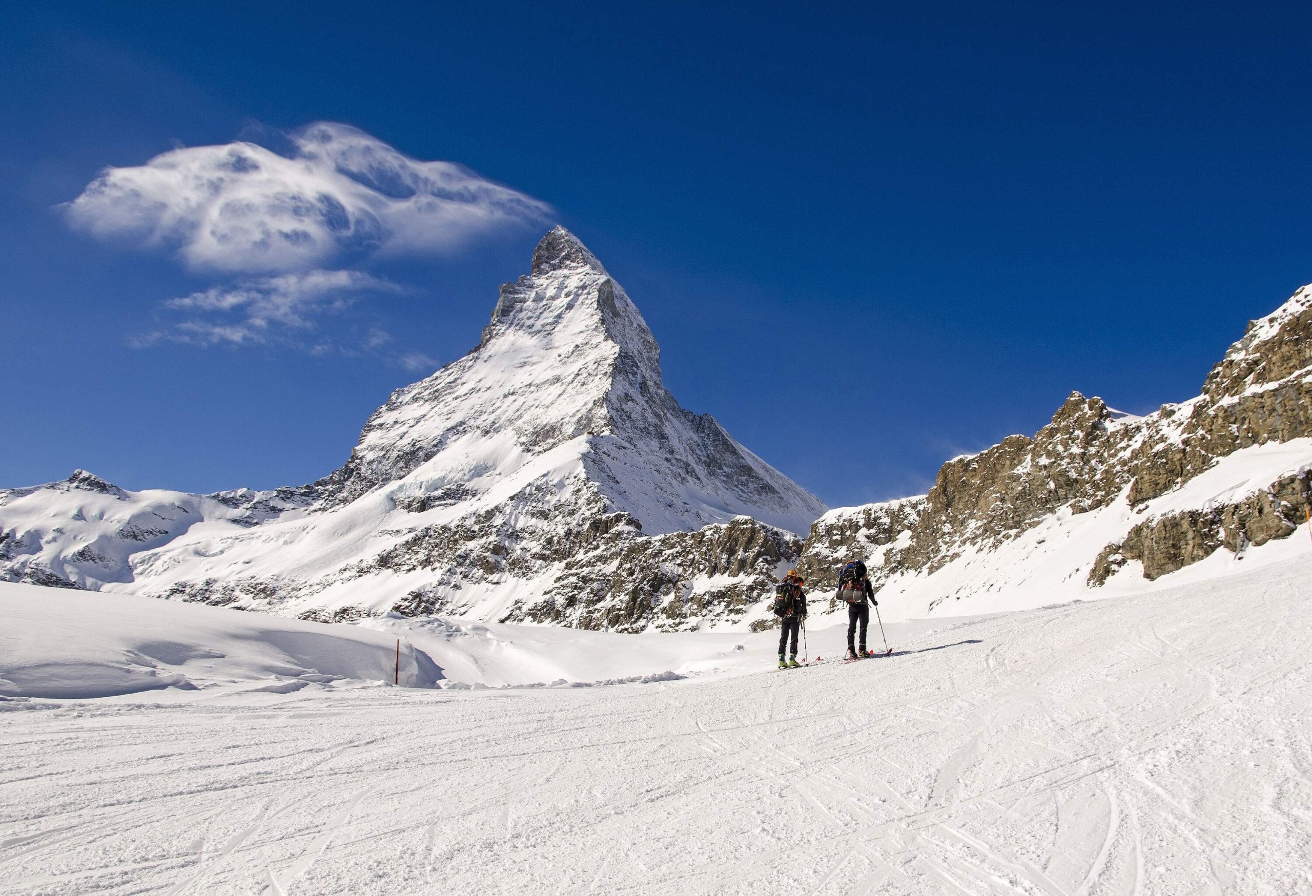 Two skiers standing on deep snow land beneath a rocky mountain covered in snow.