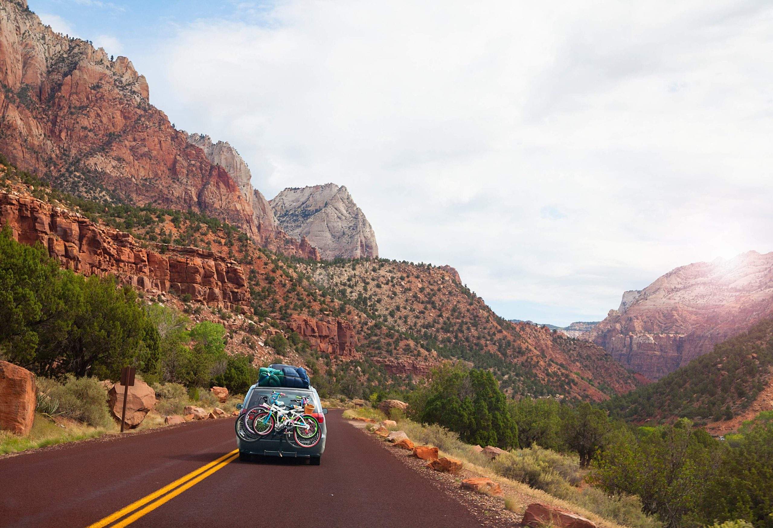 A car with bikes on the back and bags on the roof travelling down the road surrounded by sandstone mountains.