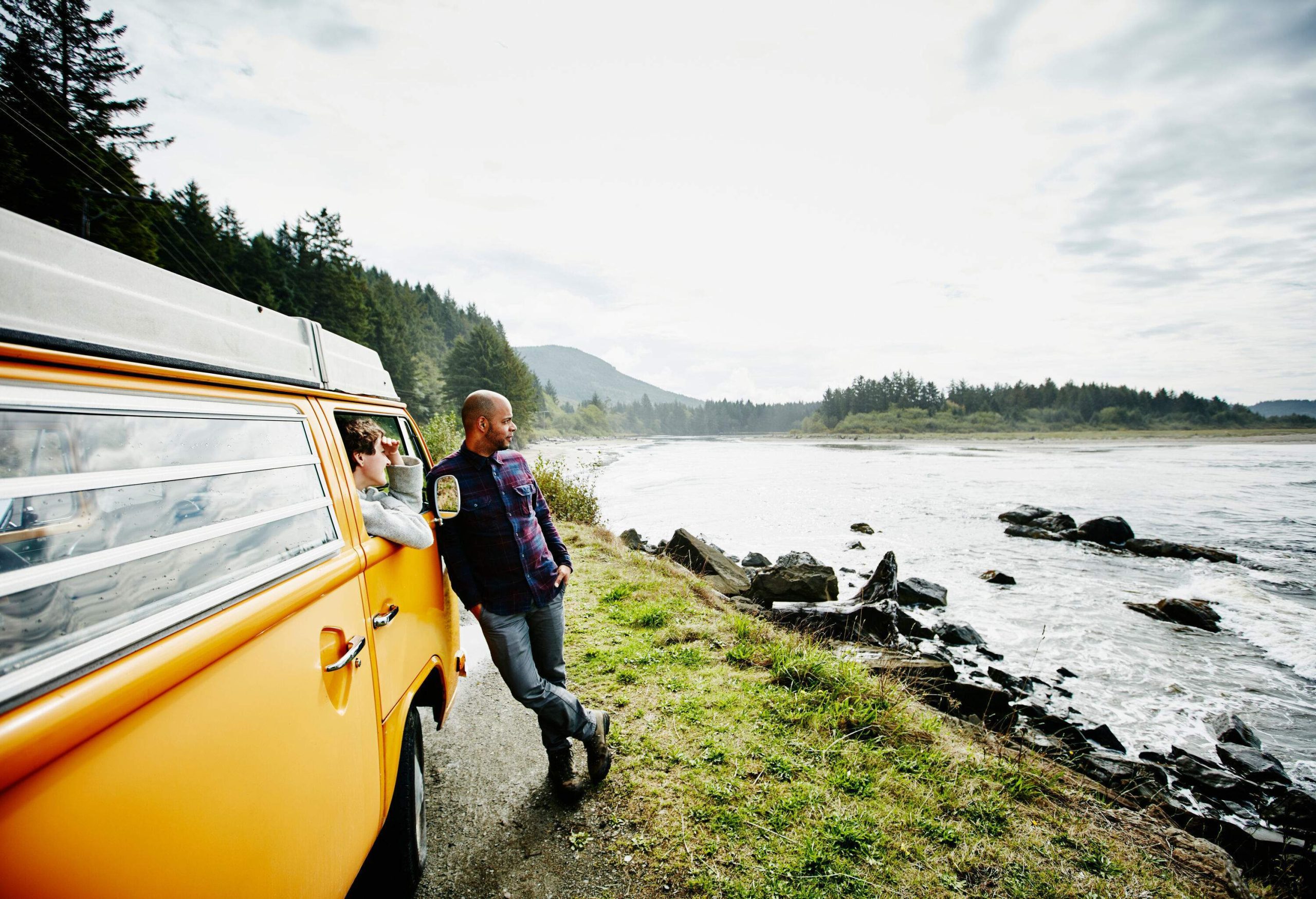 A woman peering out the window and a man leaning against the side of a van, gazing out into the ocean.