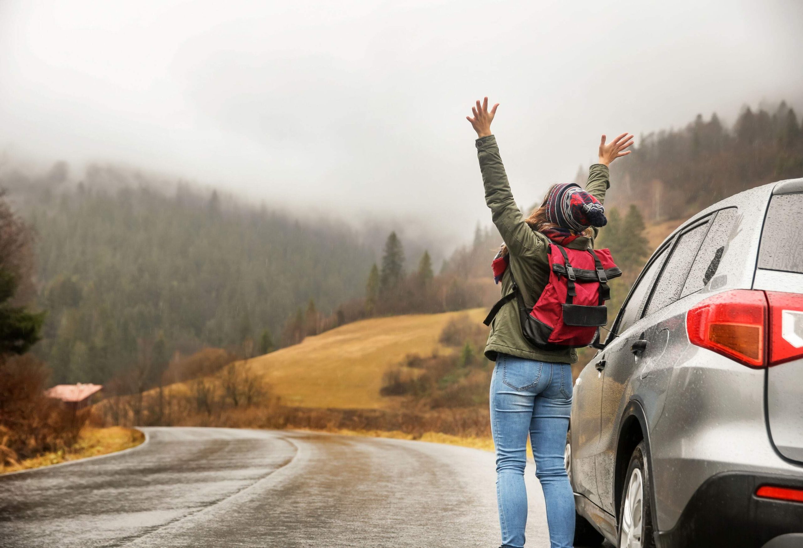 A woman in a casual winter outfit standing on the side of a wet road next to a car, looking up to the grey skies with arms raised.