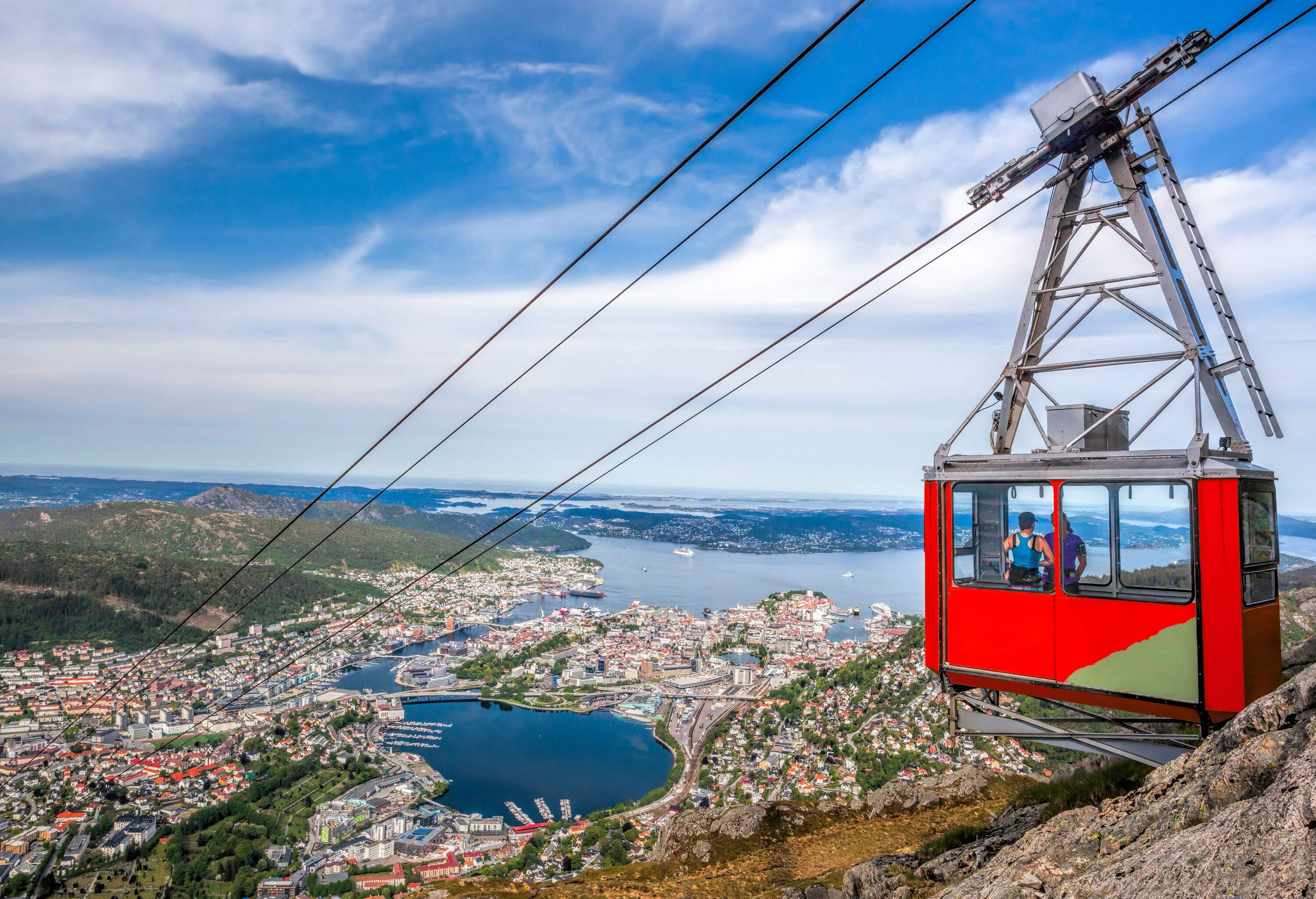 A cable car transporting two passengers descending a hillside with a populated coastal town below.