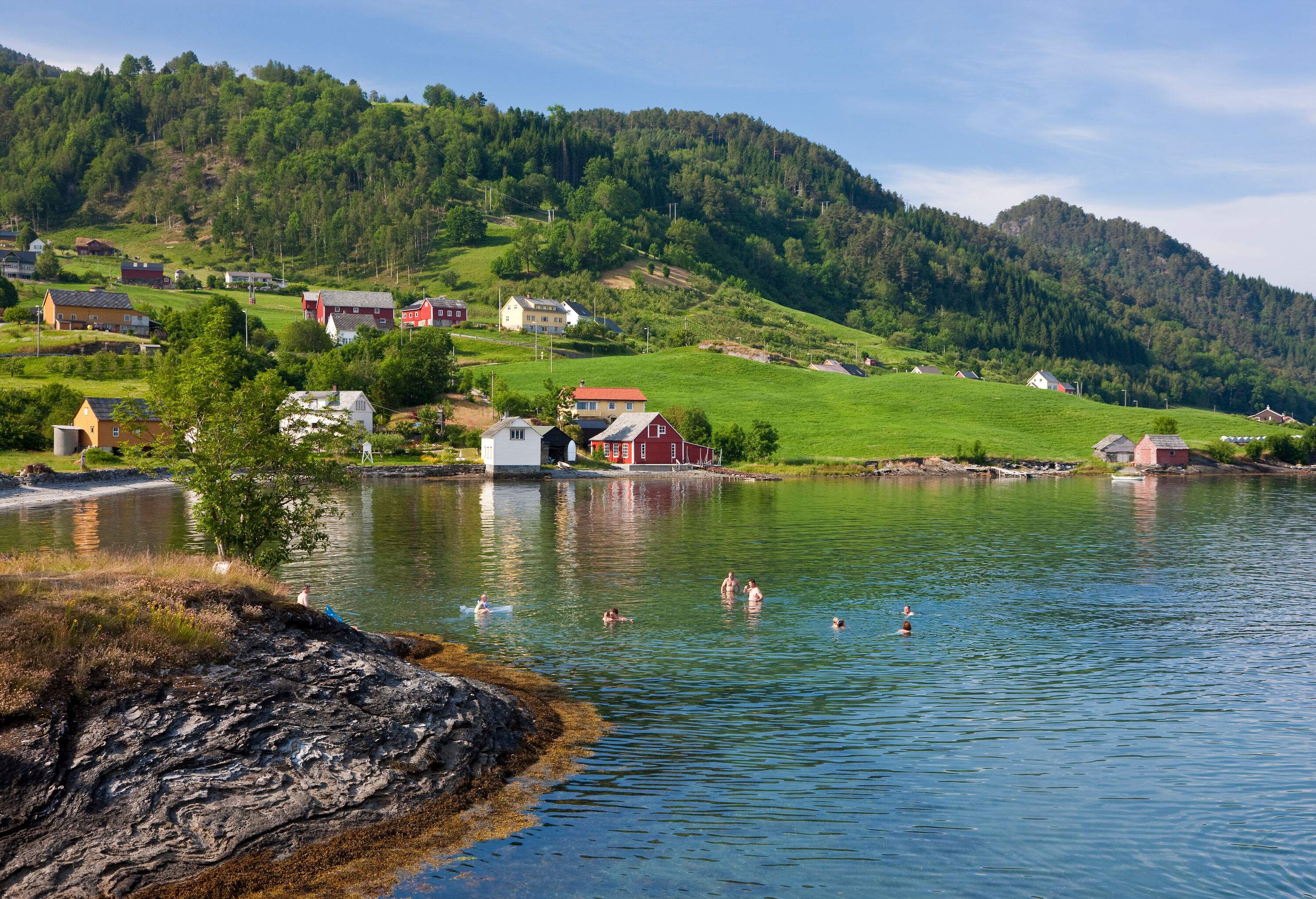 A group of people swimming in a lake along a village with houses perched on a grassy hill.