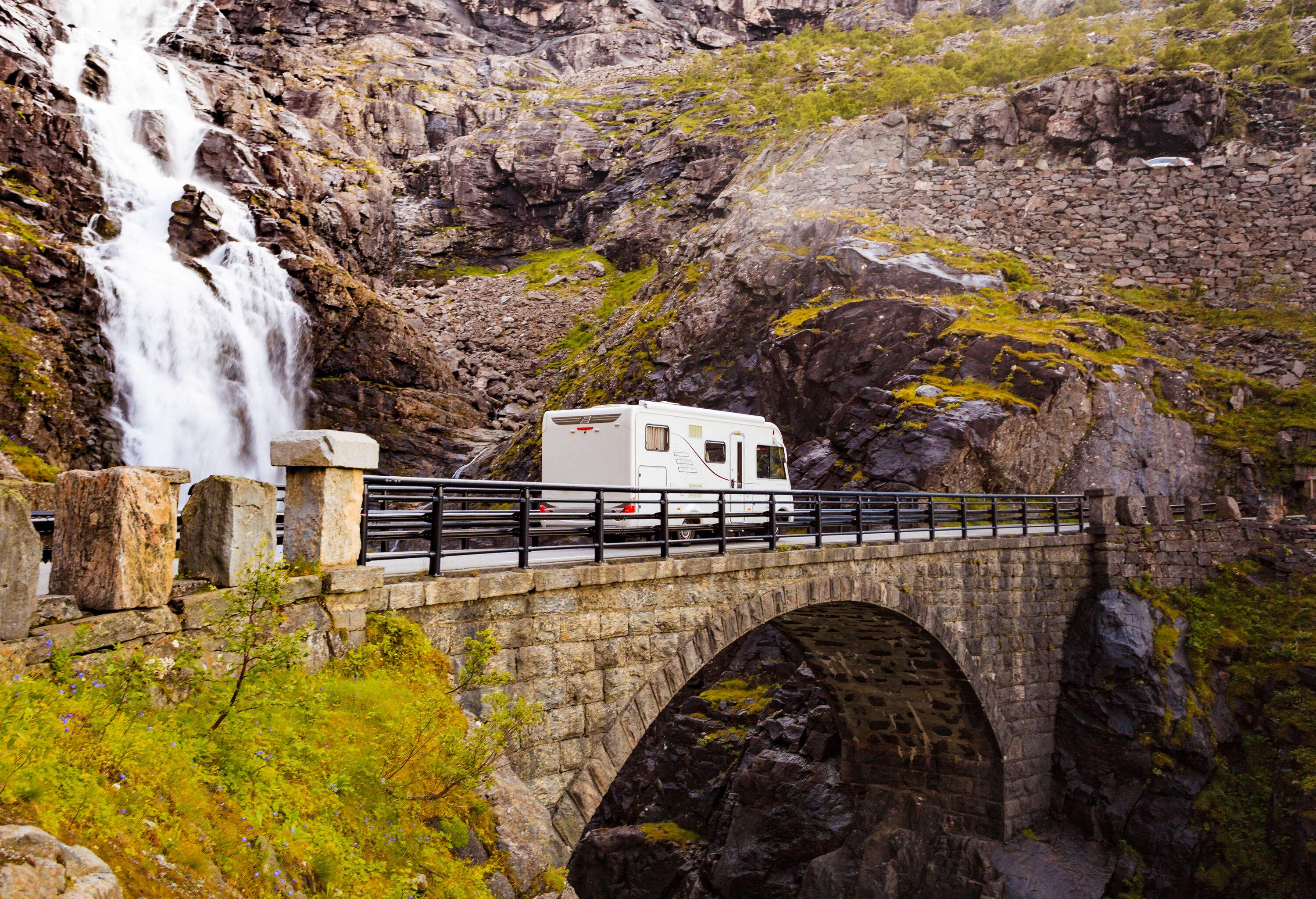 A white camper van crosses the arch bridge along a waterfall cascading through the steep.
