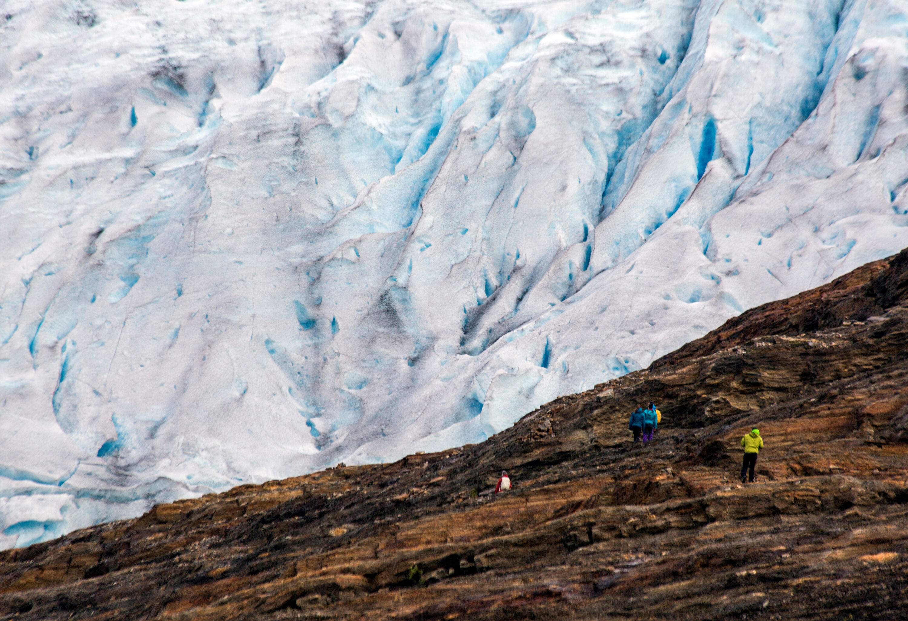 People walk on a rugged trail towards the massive ice formations of a glacier.