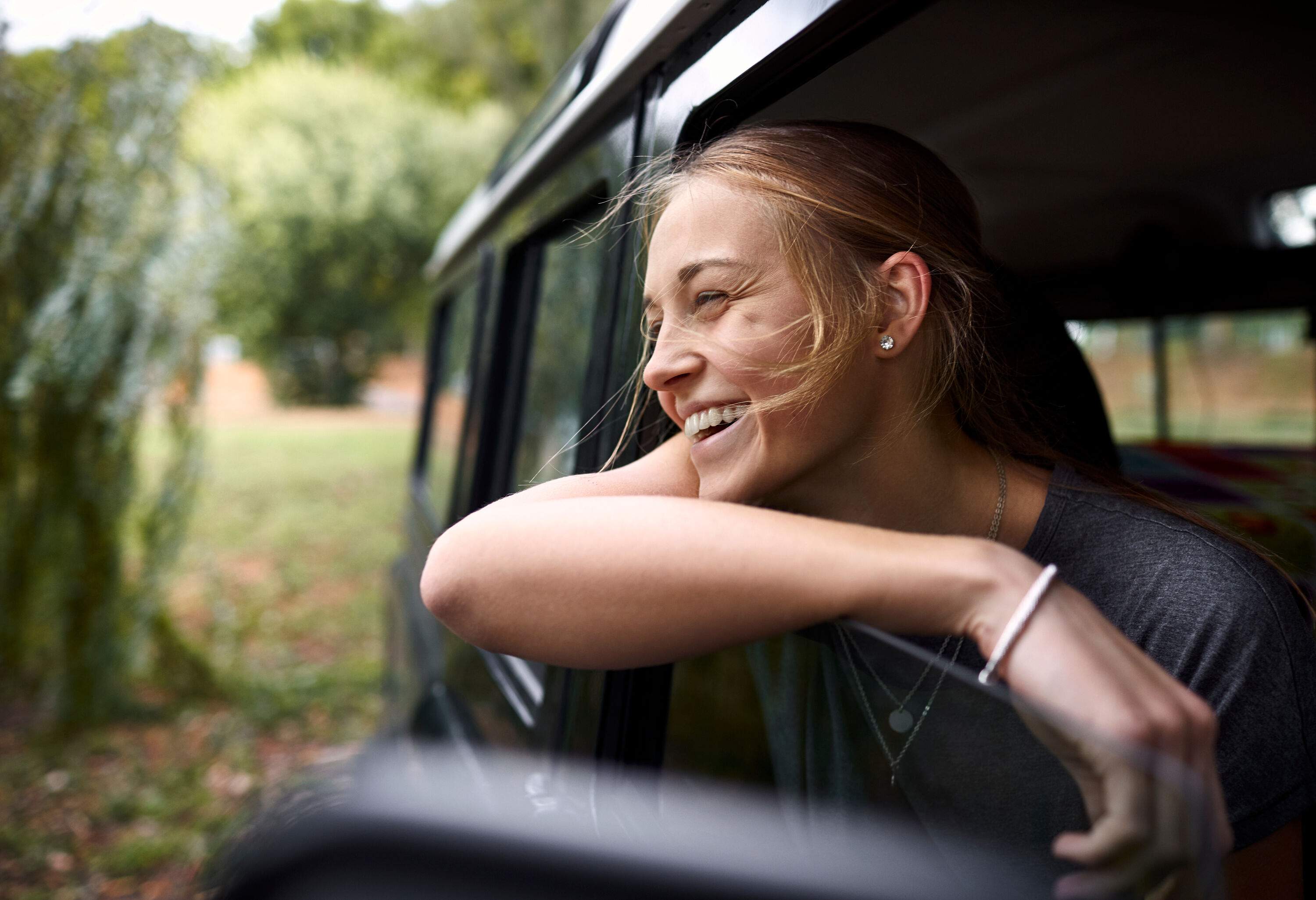 A woman smiles as she leans on the car's window while enjoying nature's breeze.