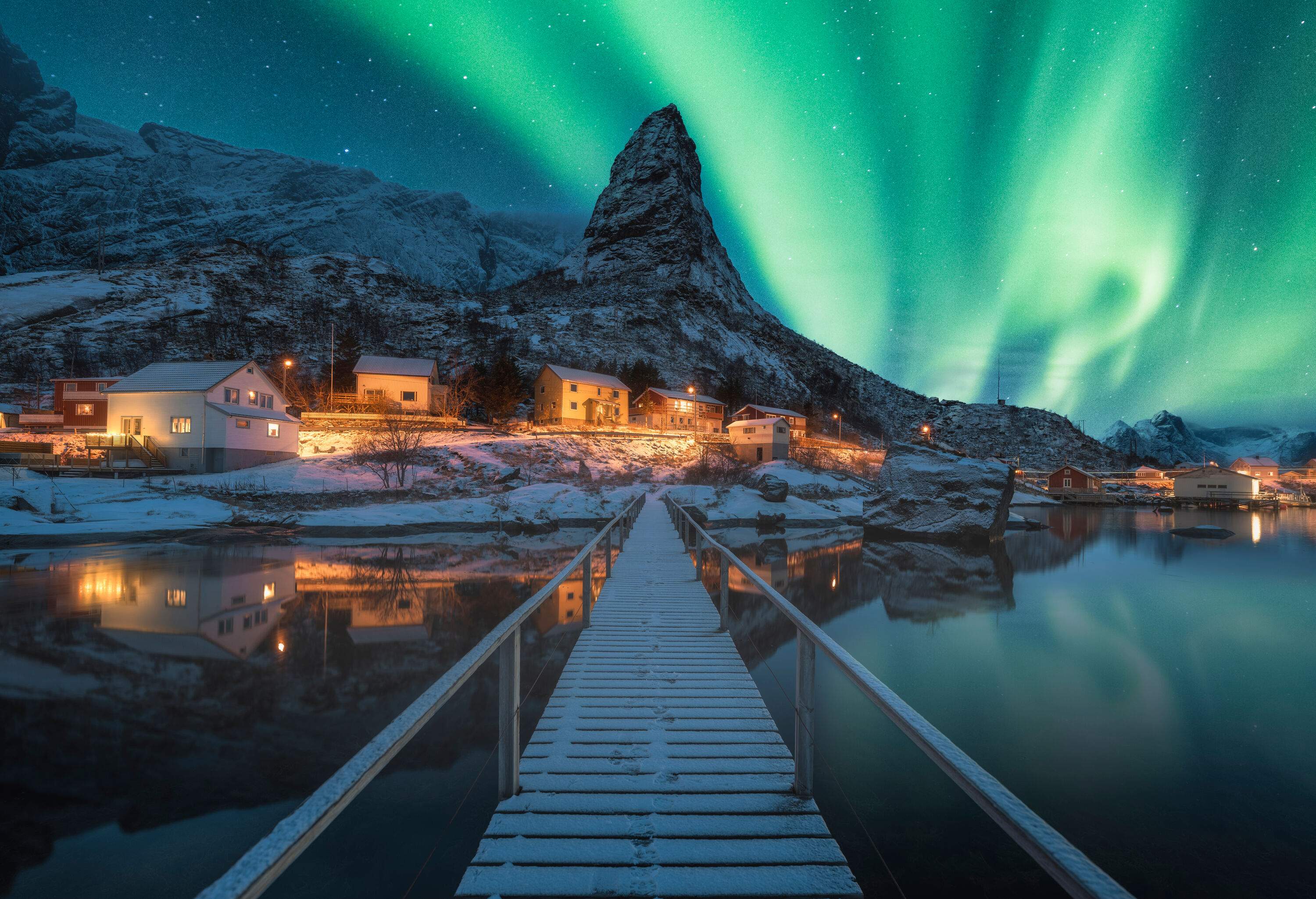 A boardwalk across the sea to a small fishing village lit up by the northern lights blazing across the sky.