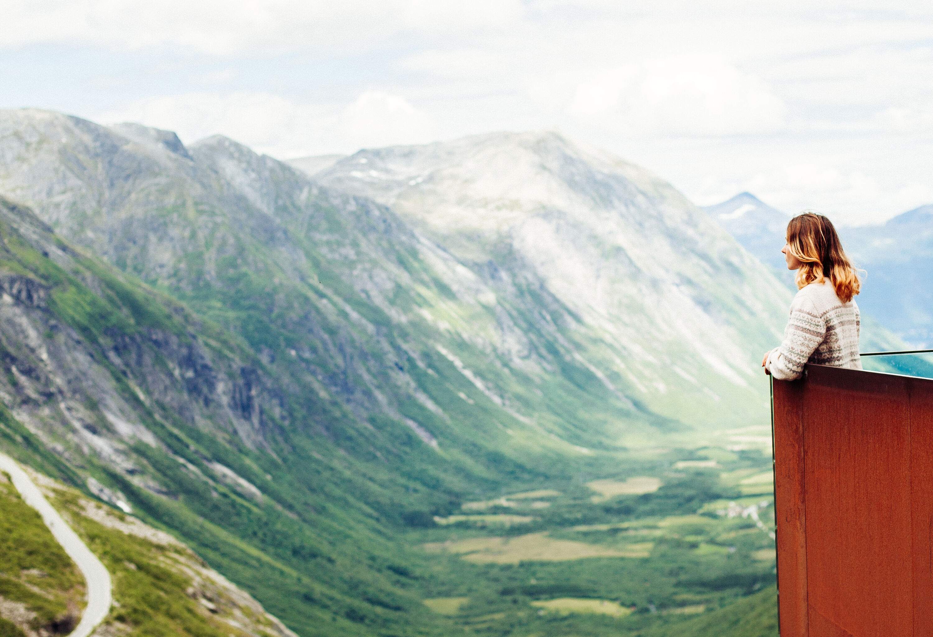 A tourist standing in a viewing platform that sits on the edge of a cliff, admiring the view over the valley and the famous winding road beneath the mountains.