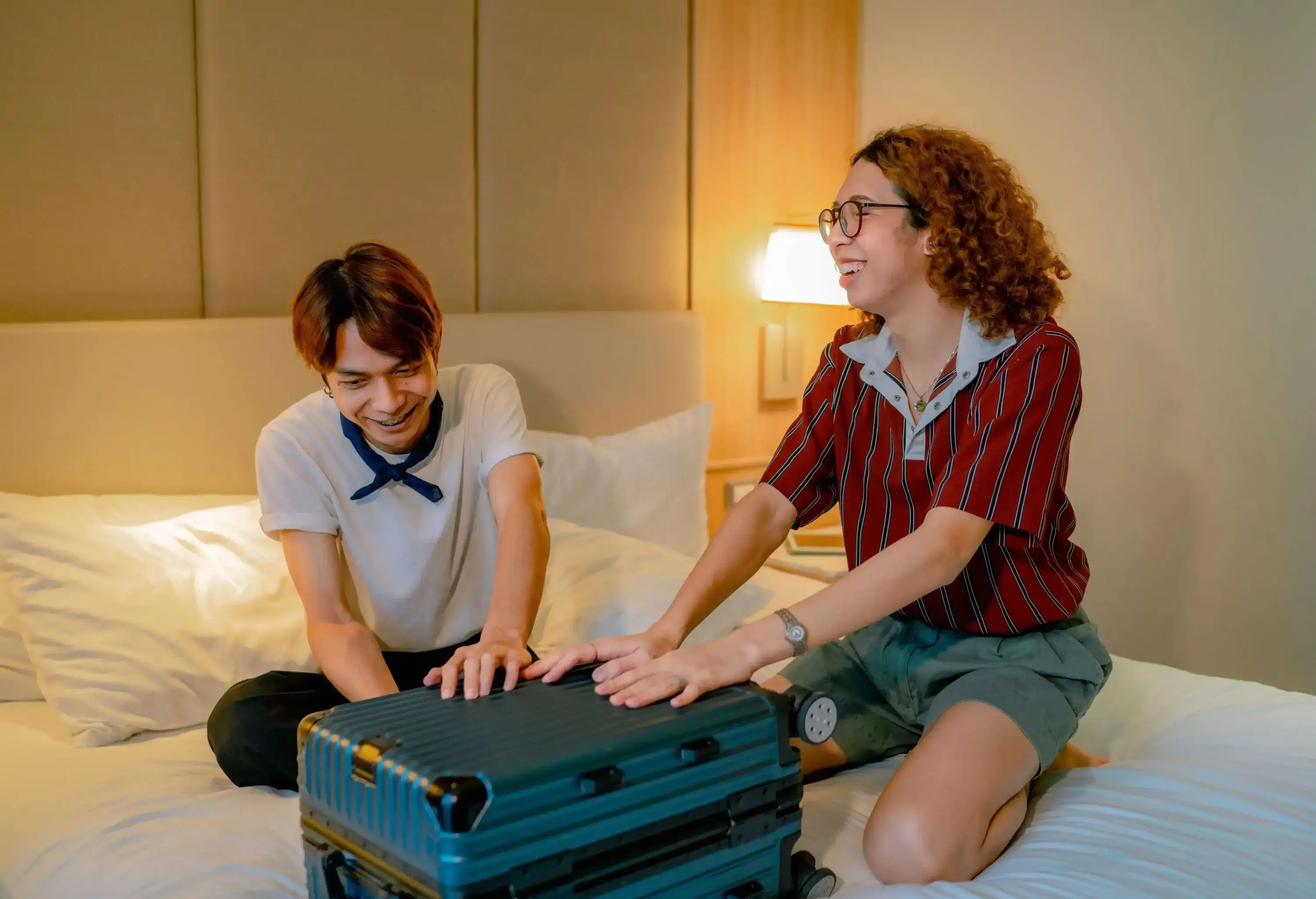 Two friends unpack their luggage in the bed in a hotel room.