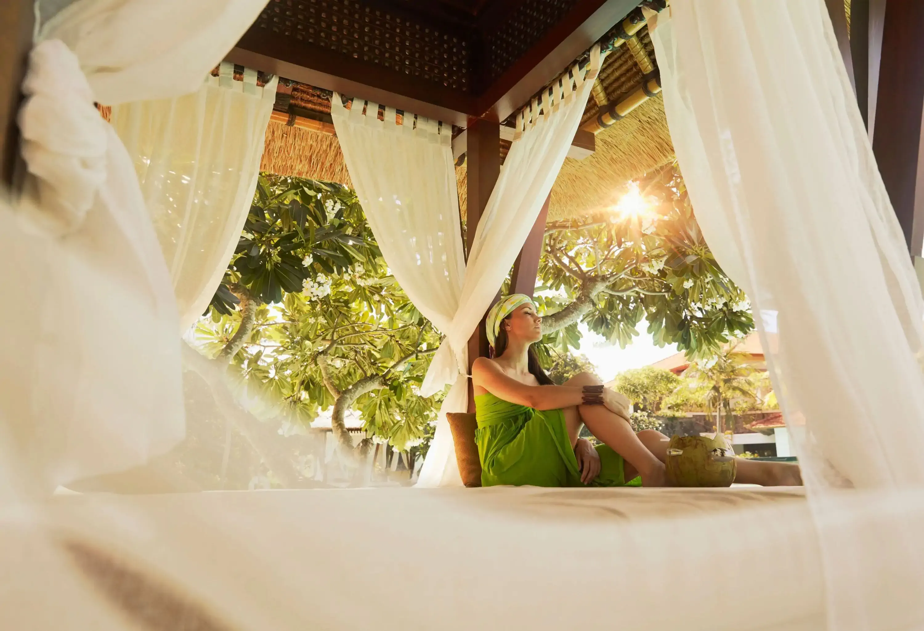 A woman in a bright green dress leaning on a canopy bed post with a coconut by her feet.