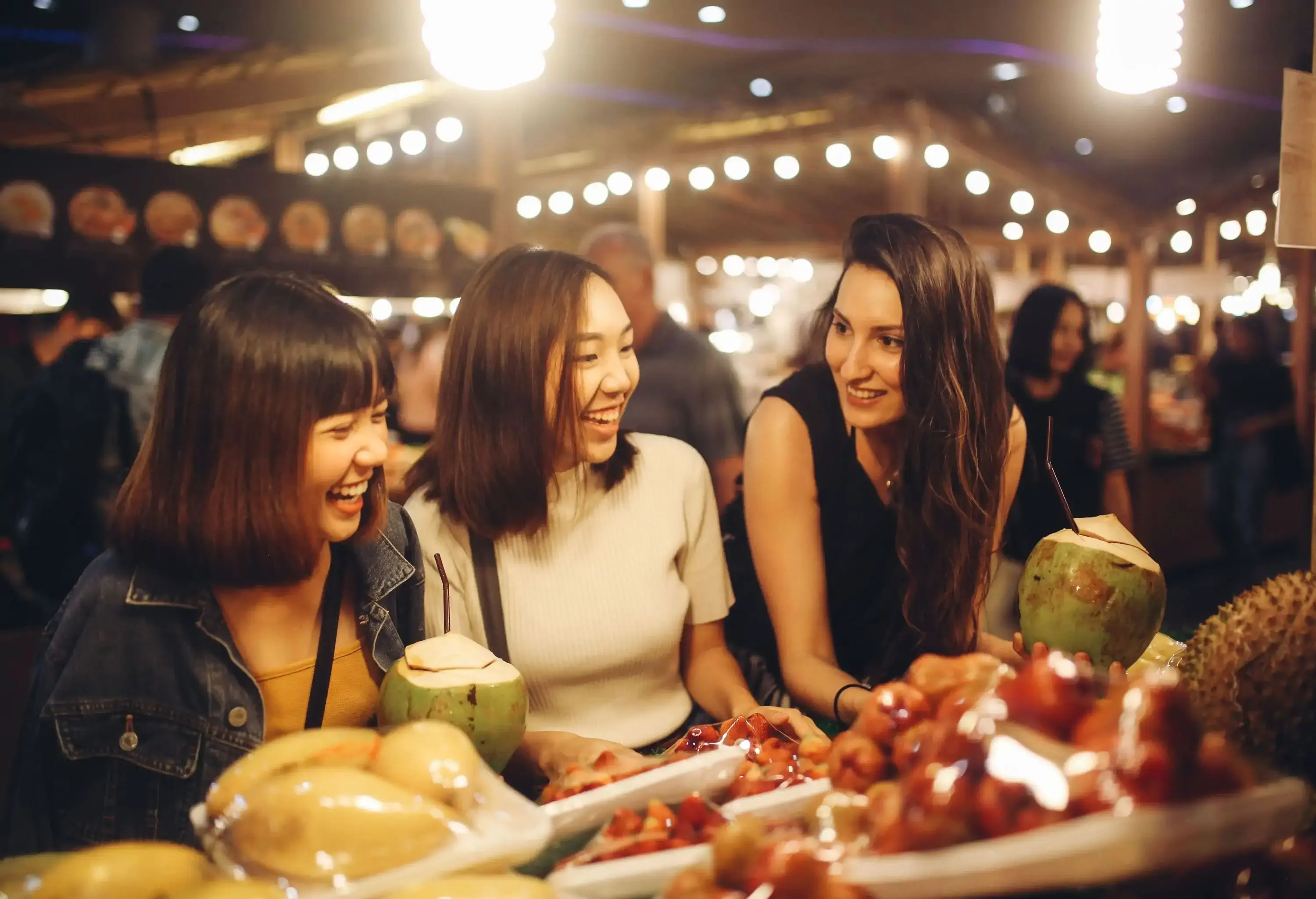 Three women smiling in front of a fruit display on a night market.