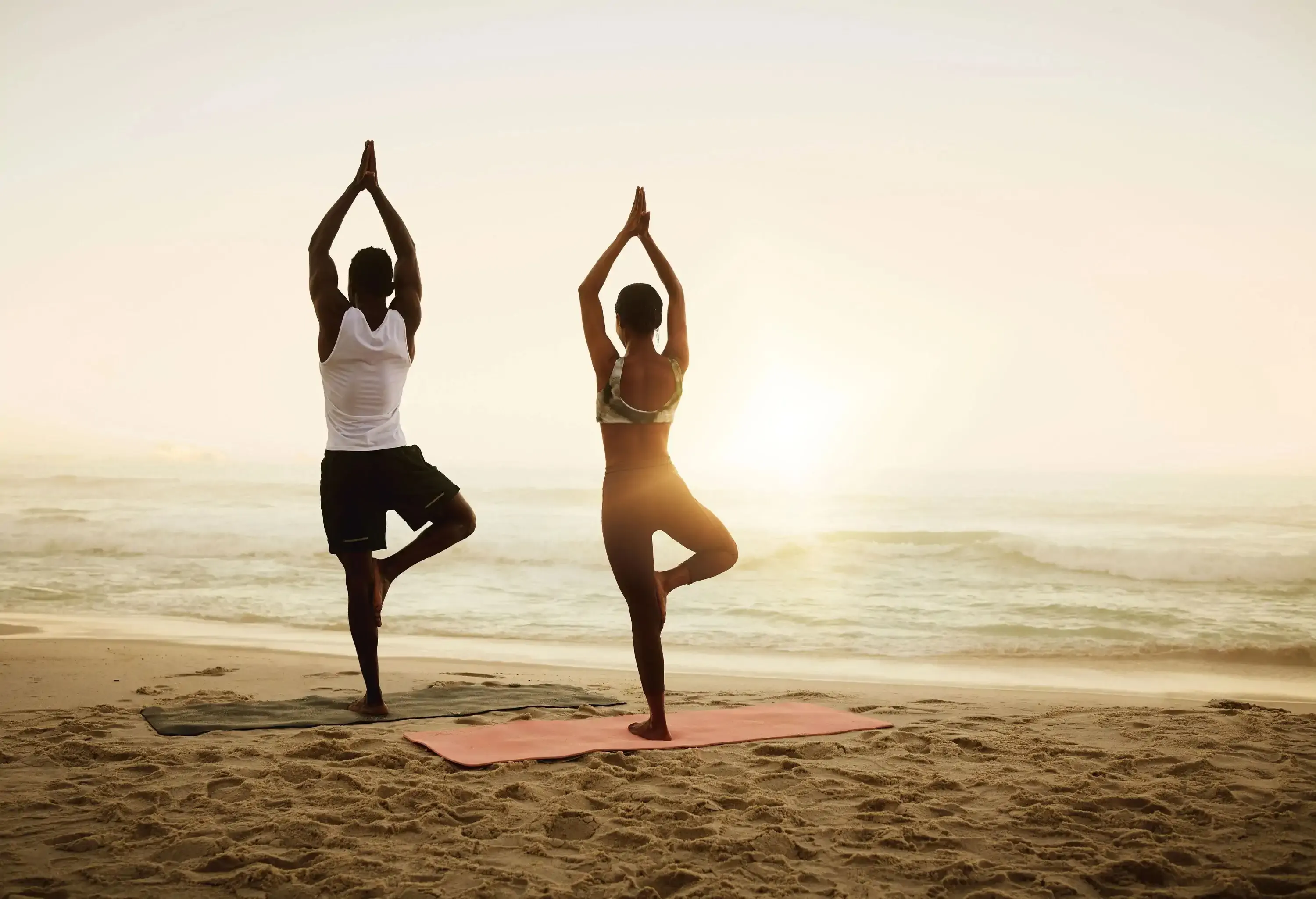A man and a woman doing yoga poses with one leg in front of a beach.