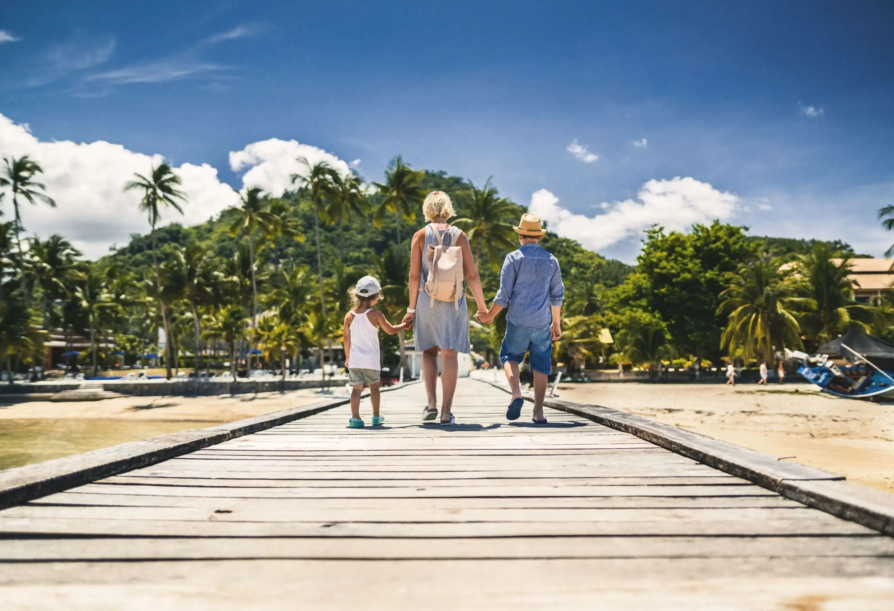 A woman and two young children holding hands as they walk toward the shore on a wooden boardwalk.