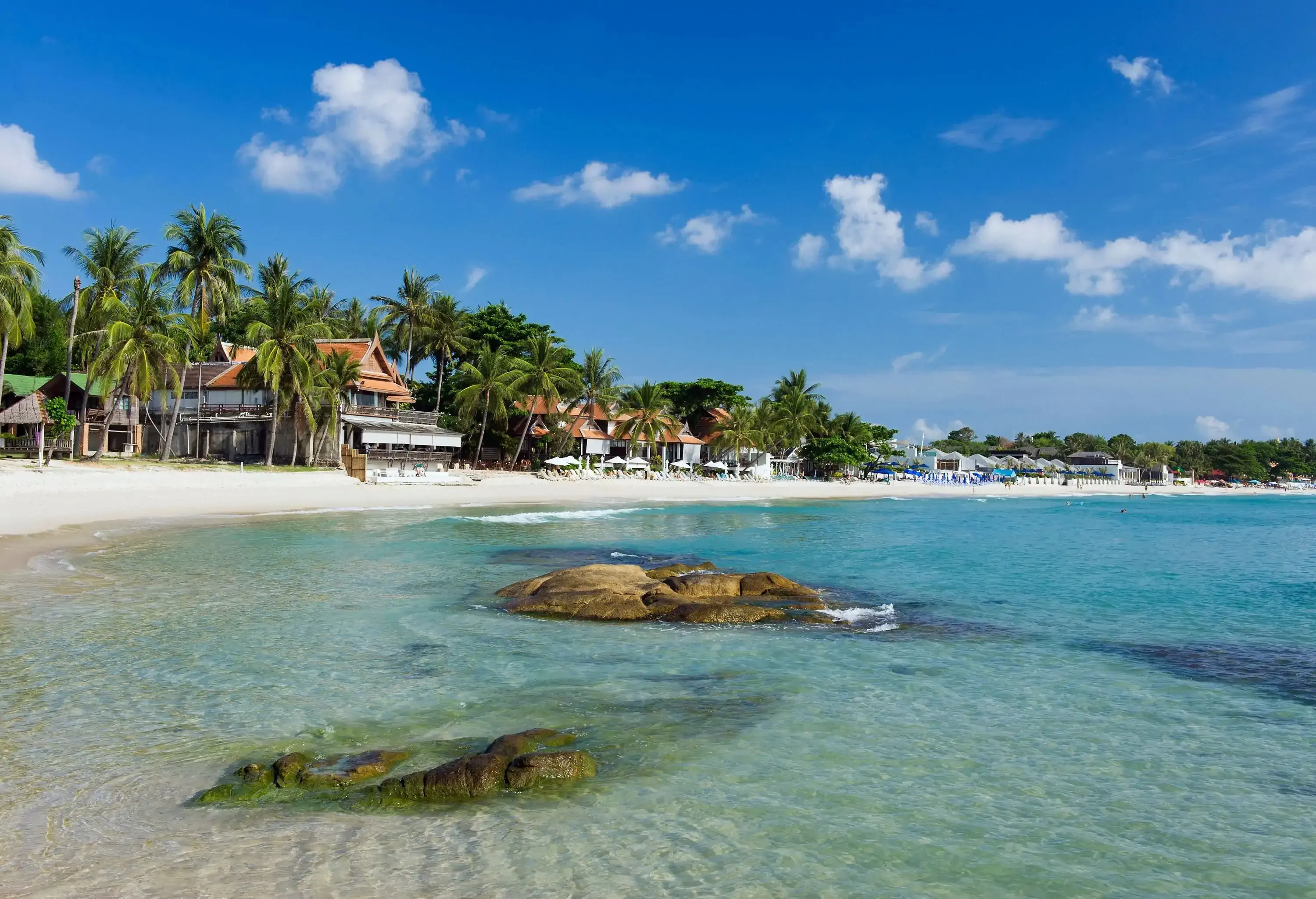 Sunken boulders in a clear sea with coconut trees and buildings lining the beach.