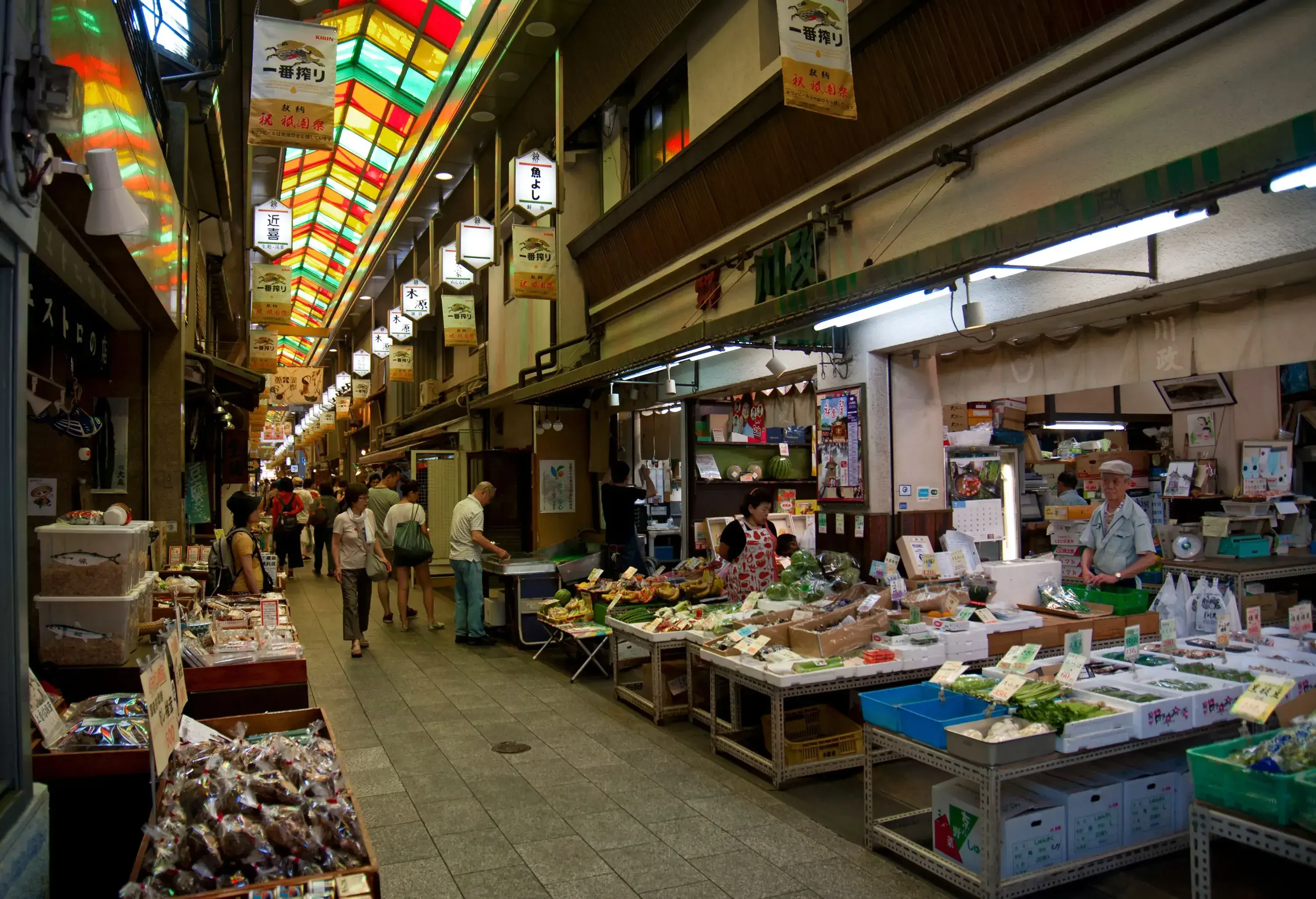 A bustling lane in a vibrant market, illuminated by skylights, is filled with people navigating through a clean environment, showcasing a diverse array of merchandise in lively stalls.