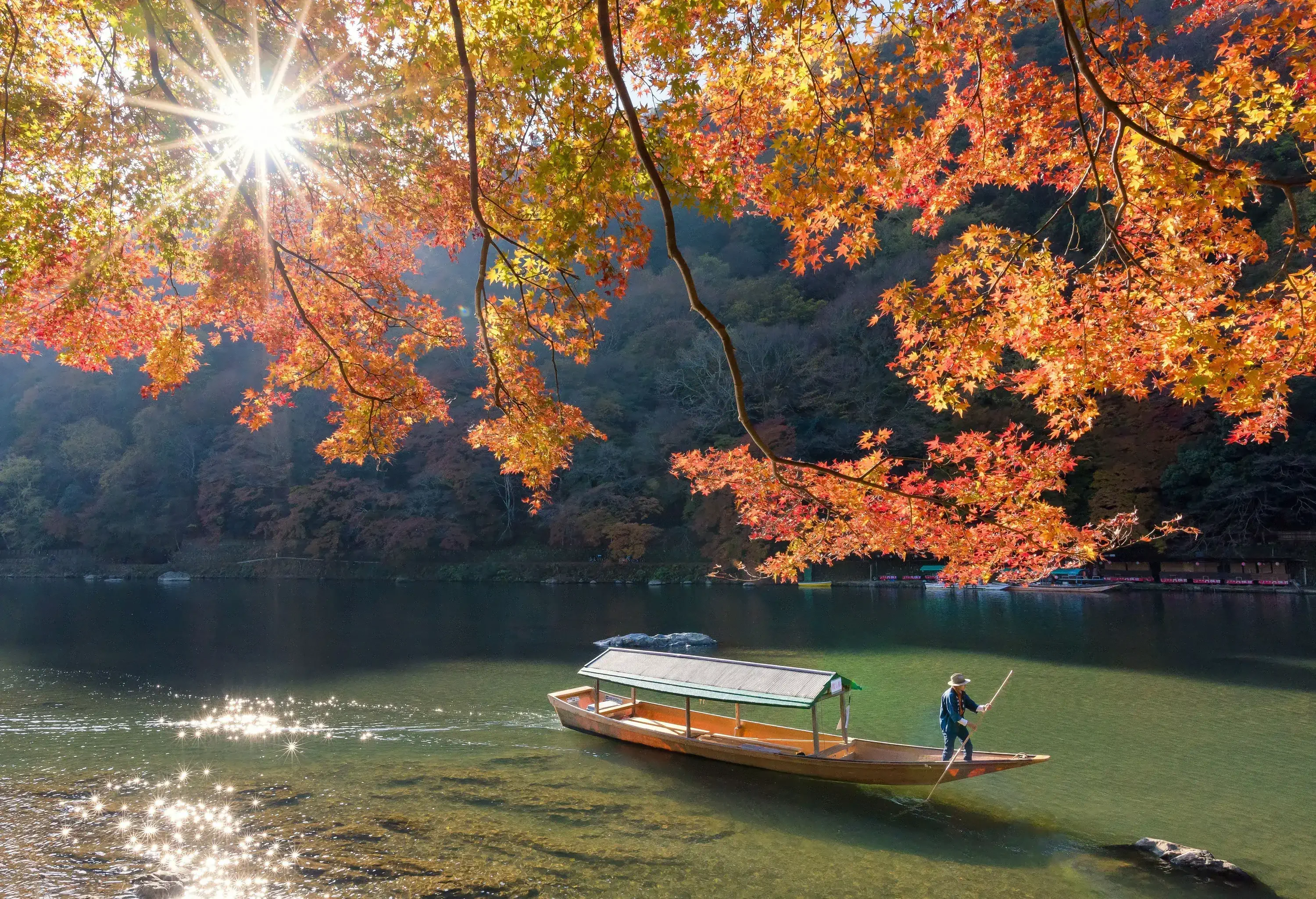 A man wearing a hat in his small boat paddling over a lake with clear waters surrounded by trees.