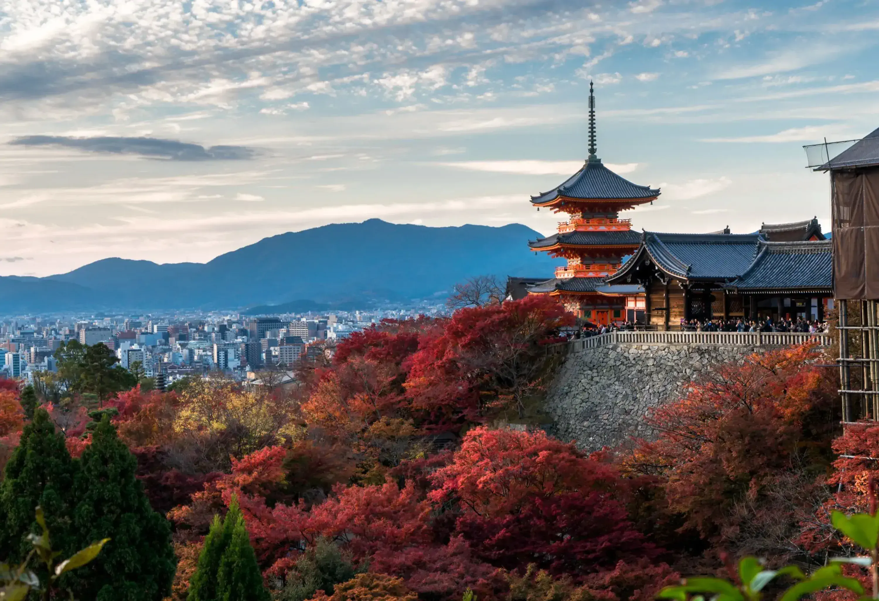 A wooden Japanese temple and pagoda surrounded by autumn trees with city buildings in the distance.