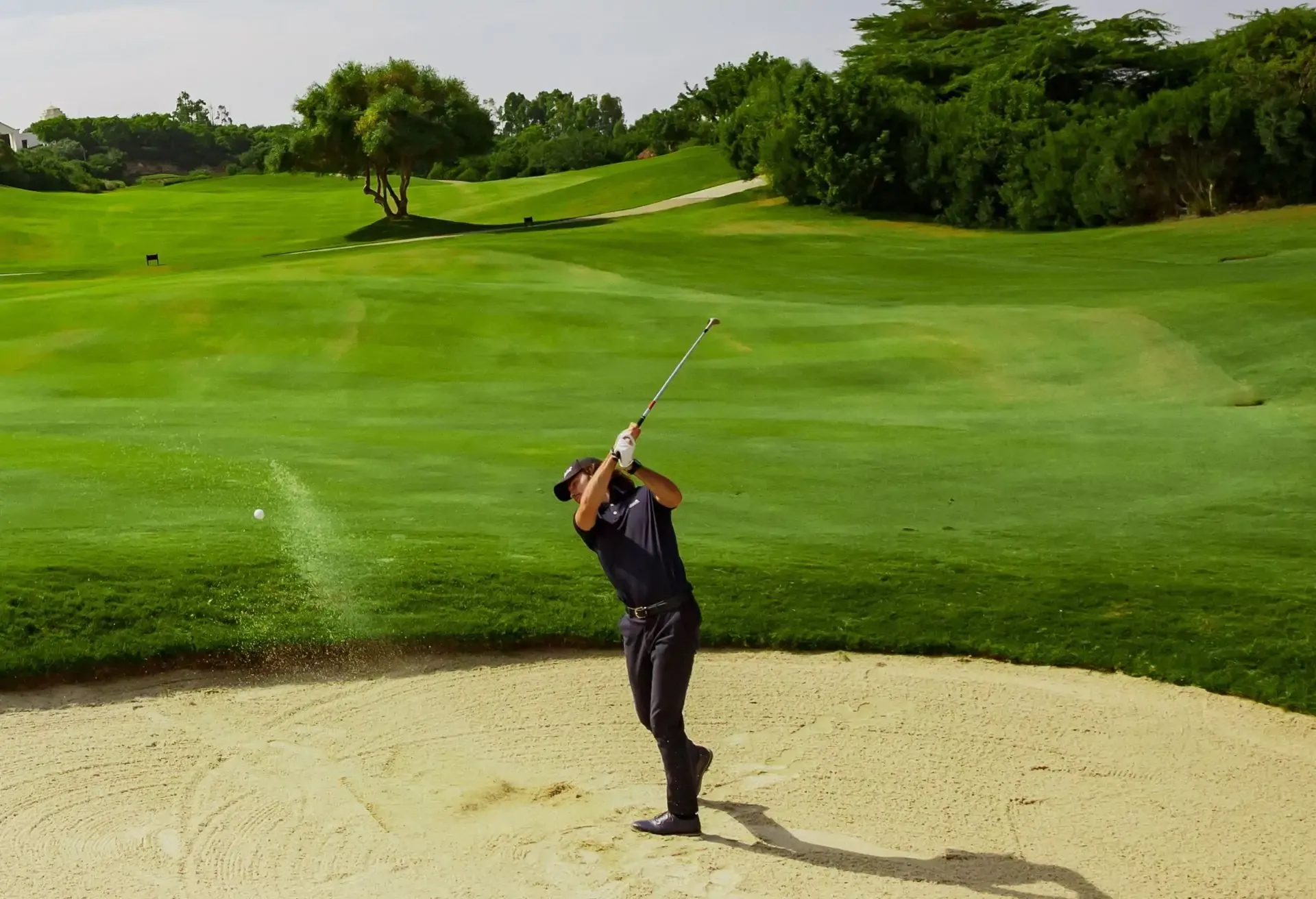 Golfer hitting a shot from a sand bunker on a lush green golf course.